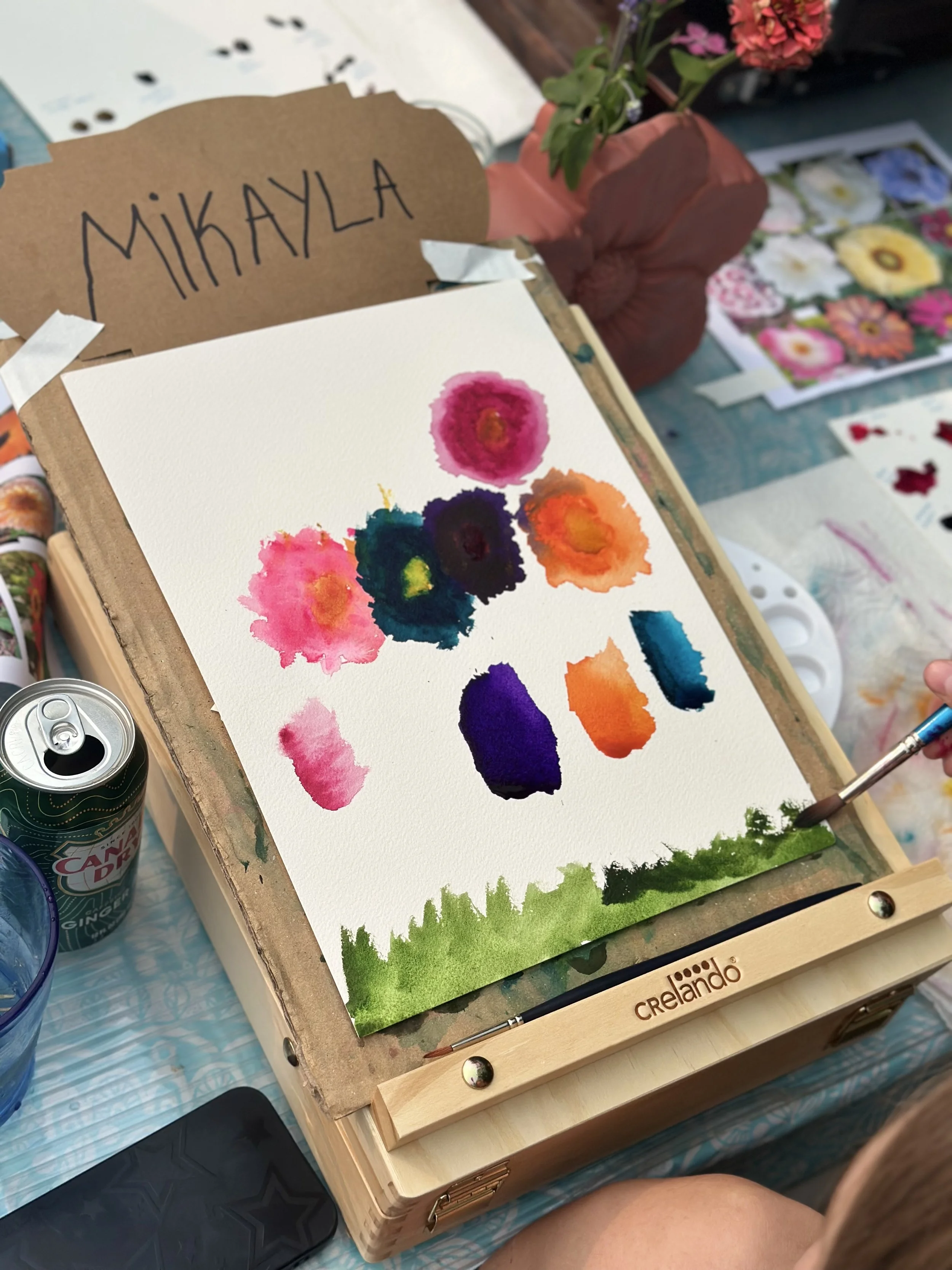 Watercolor painting of colorful flowers on white paper, with green grass at the bottom, on a wooden art box with the name 'Crelando'.