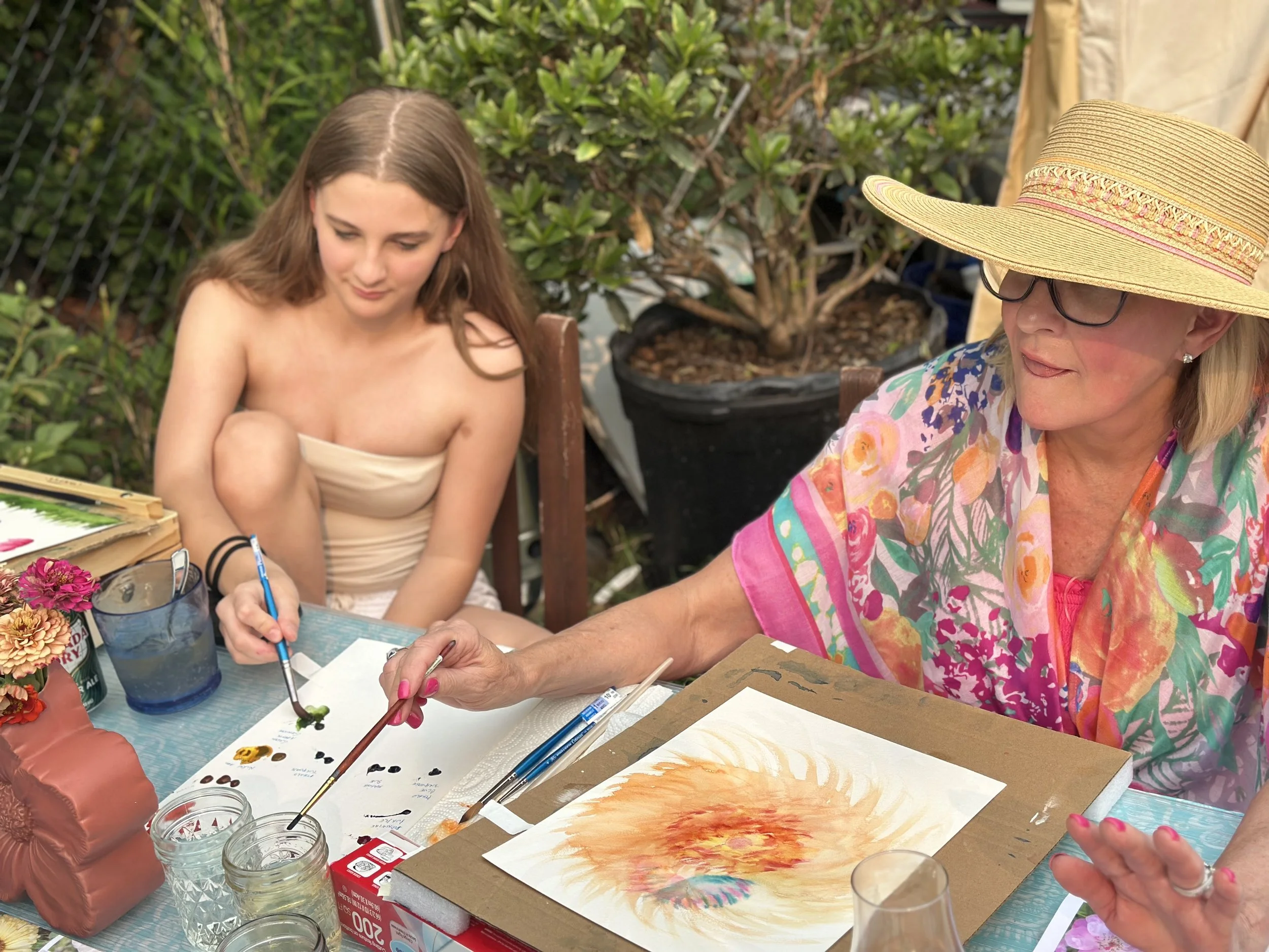 Elderly woman and young girl painting watercolor art outdoors at a table surrounded by foliage and gardening supplies.
