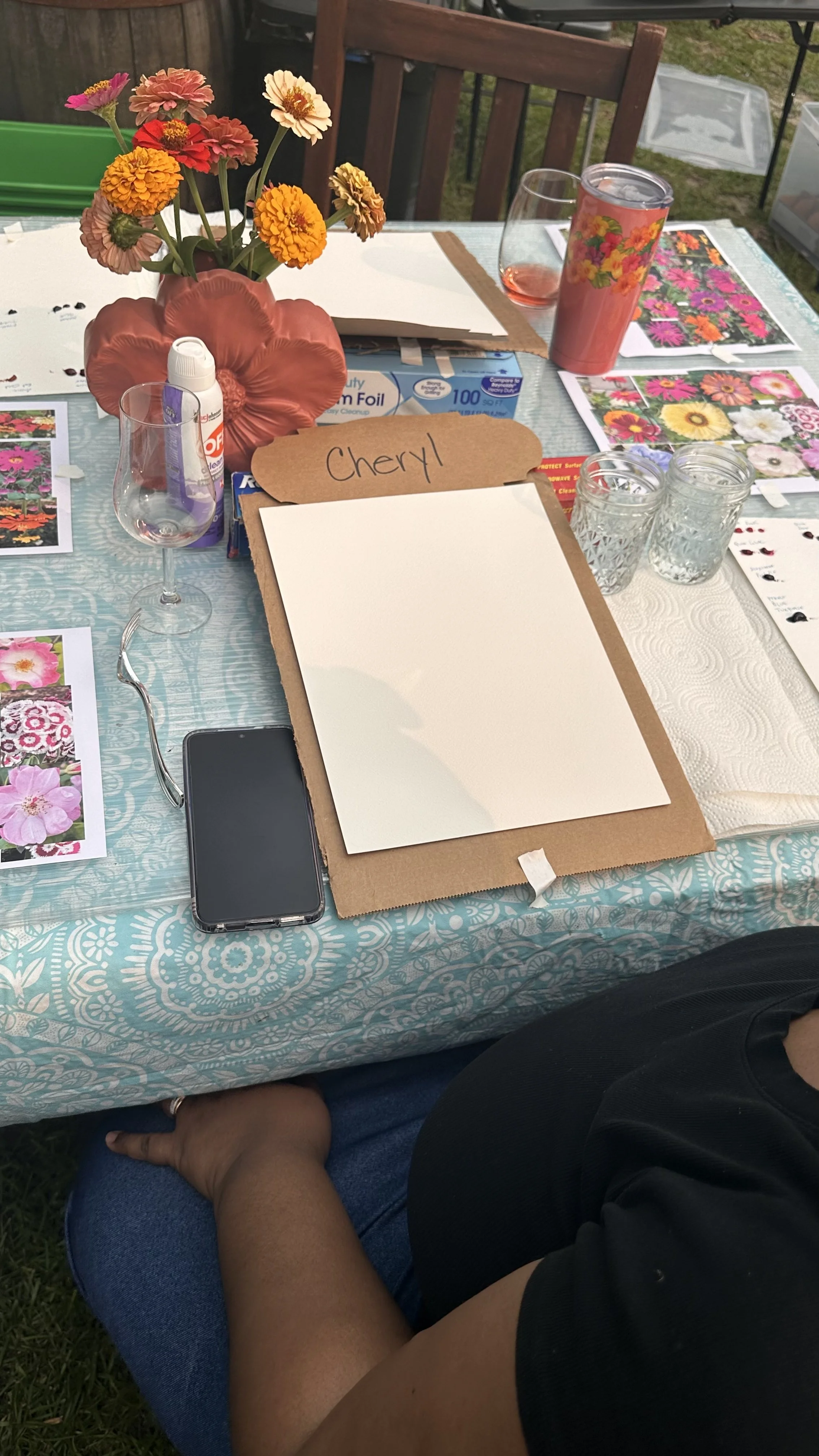 Table set outdoors with a floral centerpiece, geneology photos, a clipboard with a blank sheet and a label that reads 'Cheryl', a smartphone, two glasses, a can of pruning spray, and seed packets with colorful flowers.
