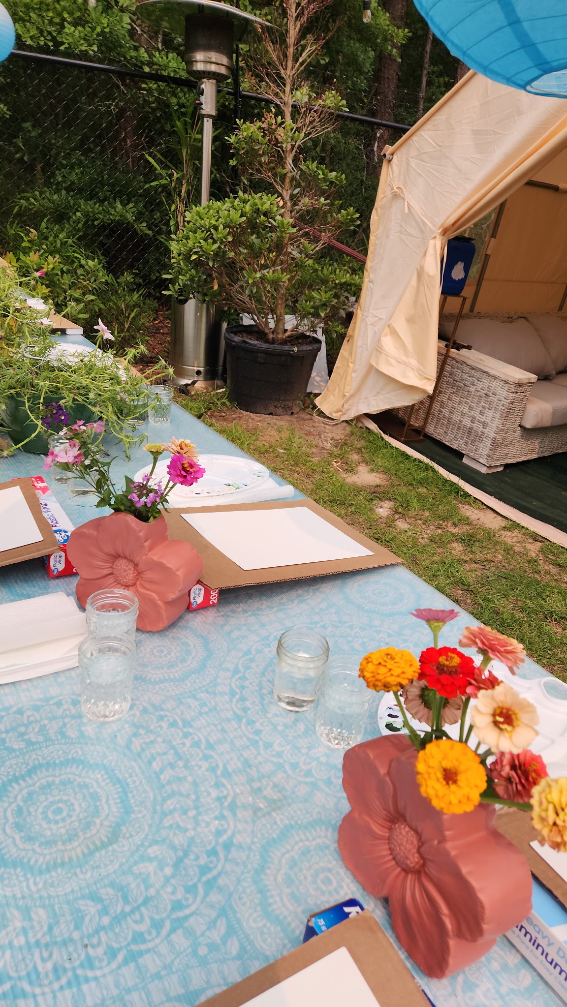 Outdoor table set with flower arrangements, glasses of water, and placemats, near a tent and lush greenery.