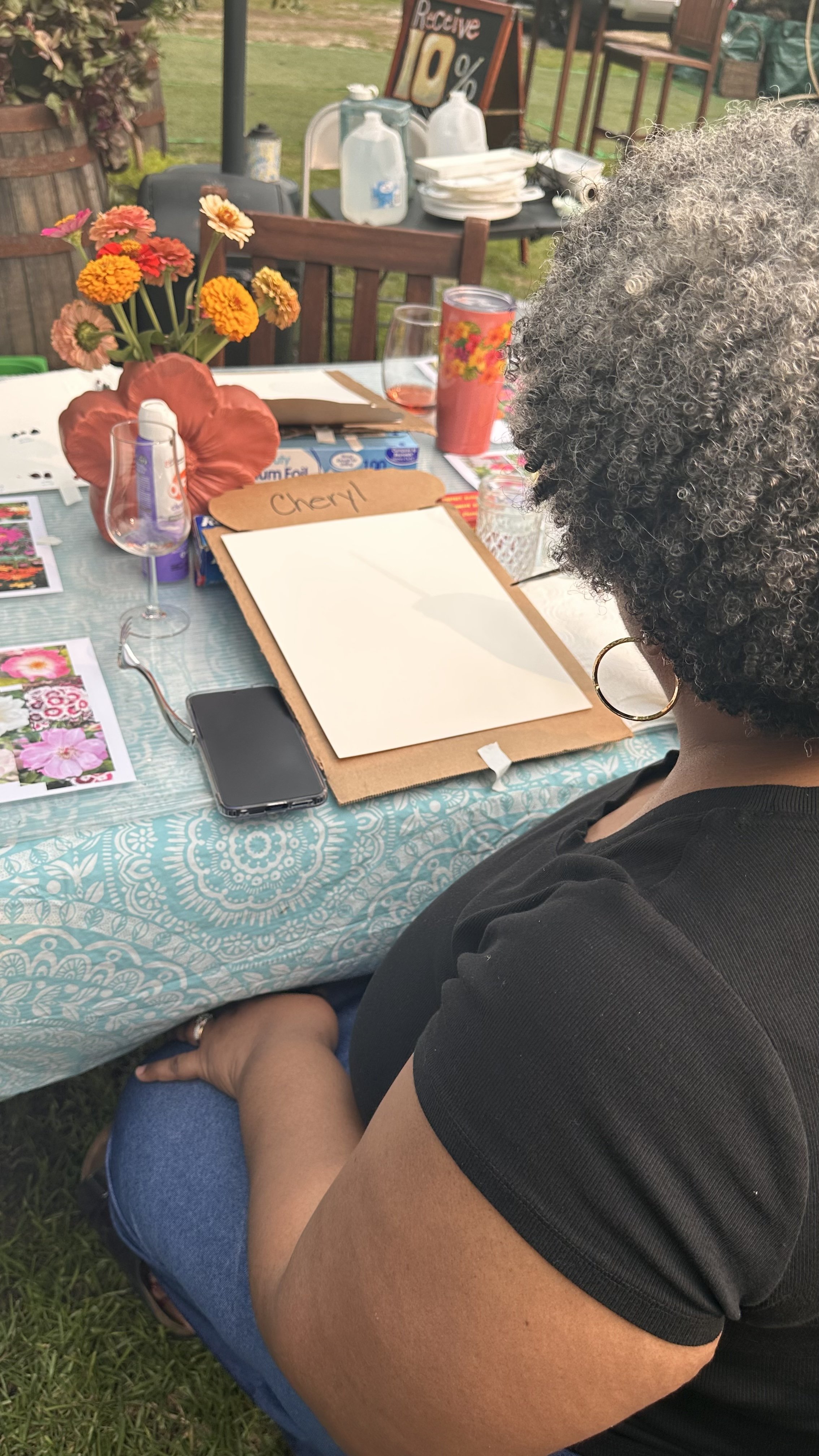 A woman with curly gray hair sitting at a decorated outdoor table with a floral tablecloth, flowers in a red vase, a menu board, and various dishware, during a gathering