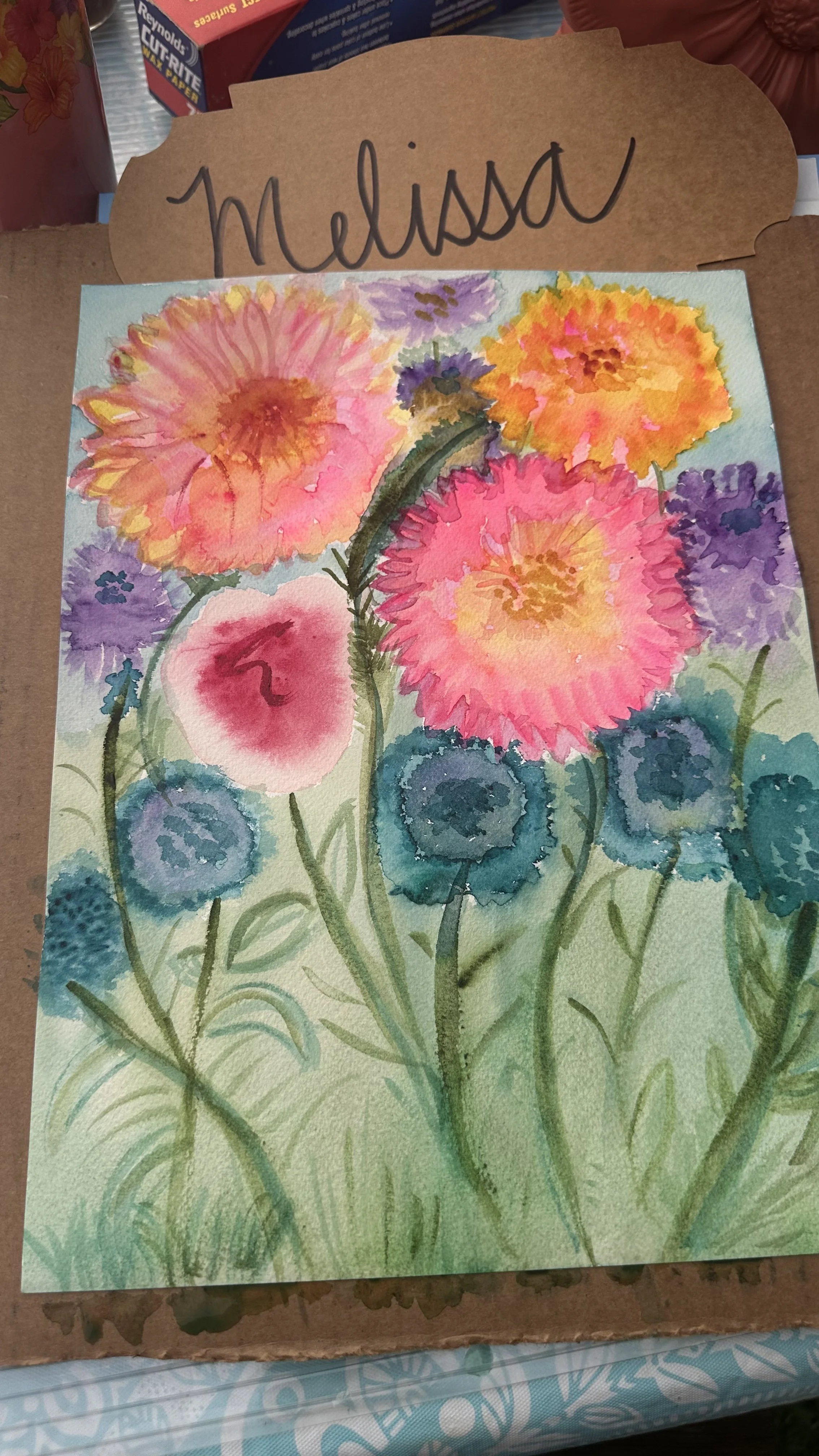 A colorful watercolor painting of various flowers in shades of pink, purple, yellow, and blue on green foliage, with a handwritten name tag that says Melissa.