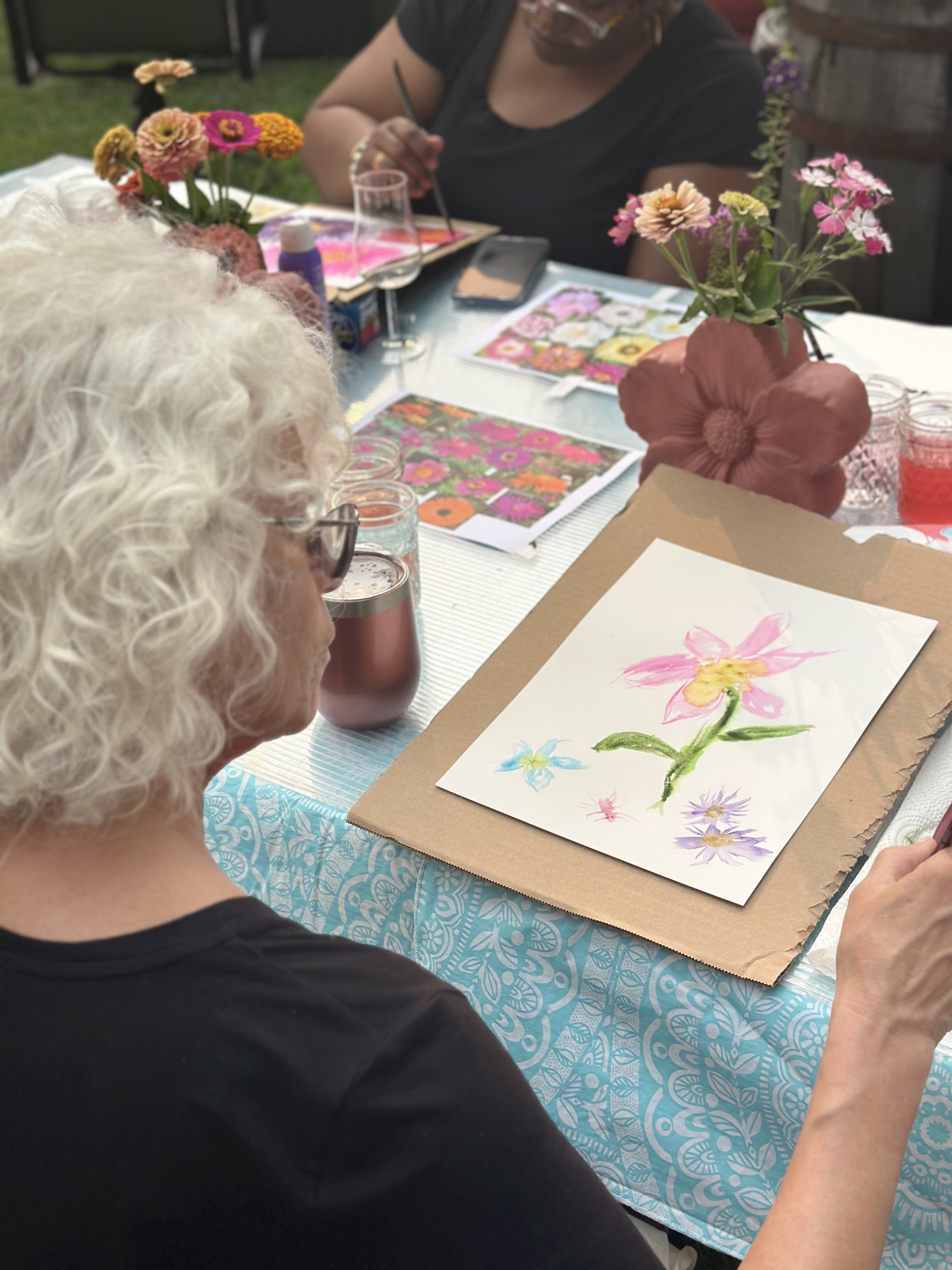 A woman with curly white hair sitting at a table, painting a colorful flower with pink, yellow, green, and purple. The table has a blue and white patterned tablecloth, with a brown paper mat underneath the paper. Other women are visible in the backgr