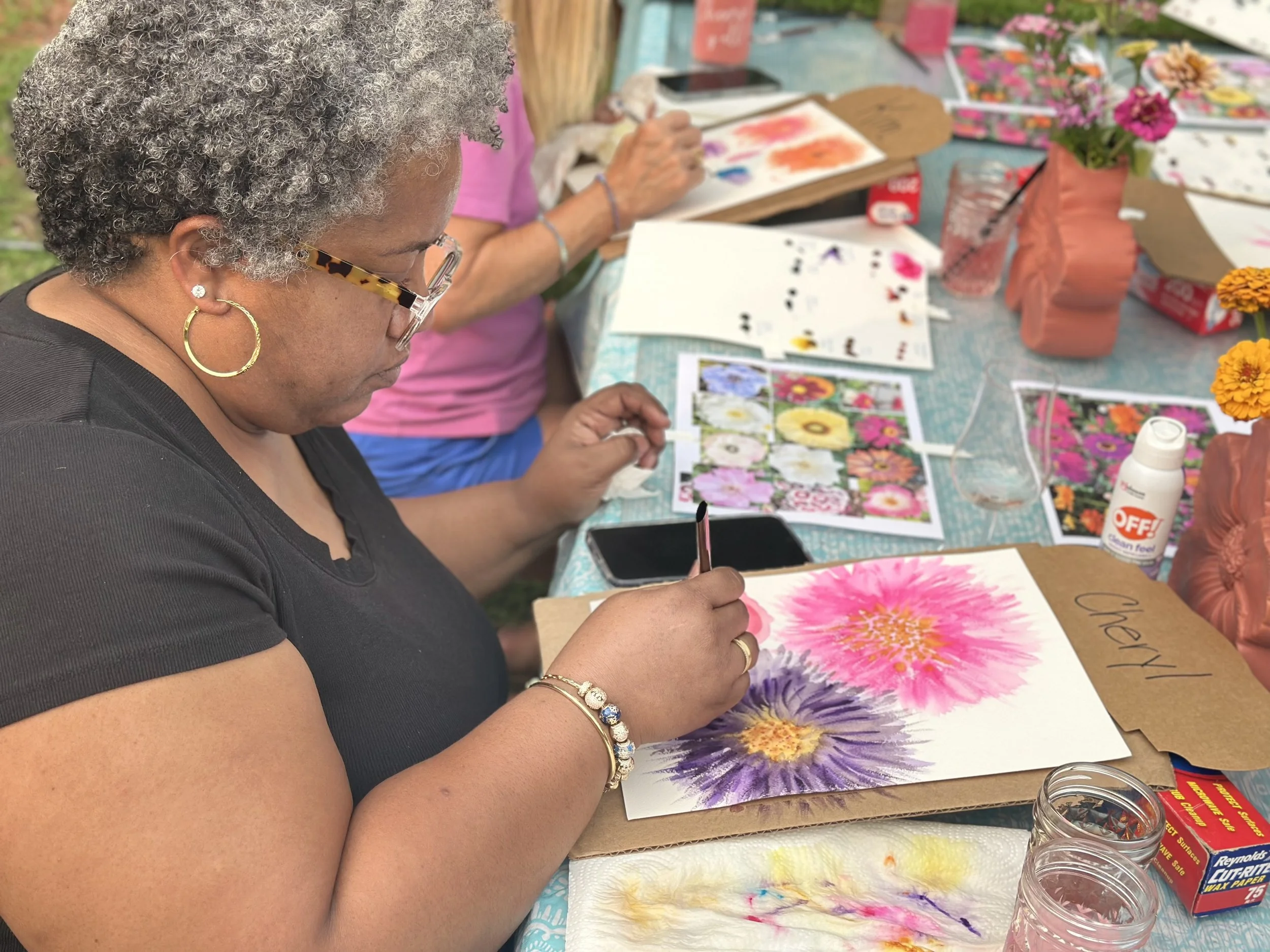 Woman with curly gray hair, wearing glasses, hoop earrings, and a black shirt, sits at a table painting a large flower watercolor with pink, purple, and yellow colors. The table is covered with art supplies, reference images of flowers, and a few sma