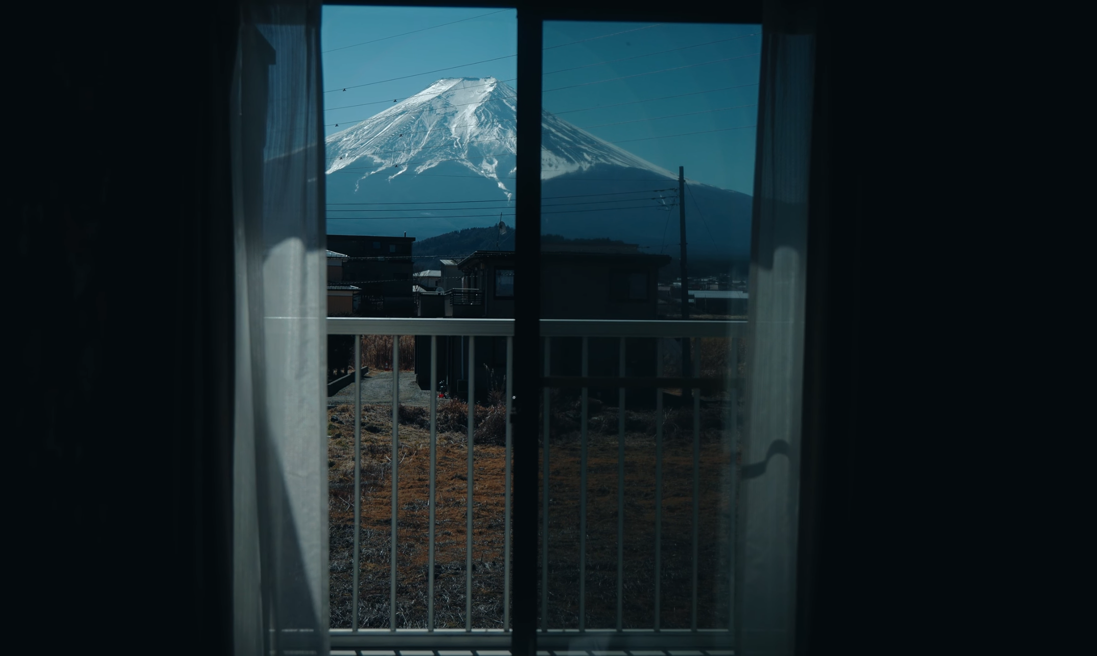 View of Mount Fuji through a sliding glass door with sheer curtains, featuring a balcony railing and suburban buildings in the foreground.