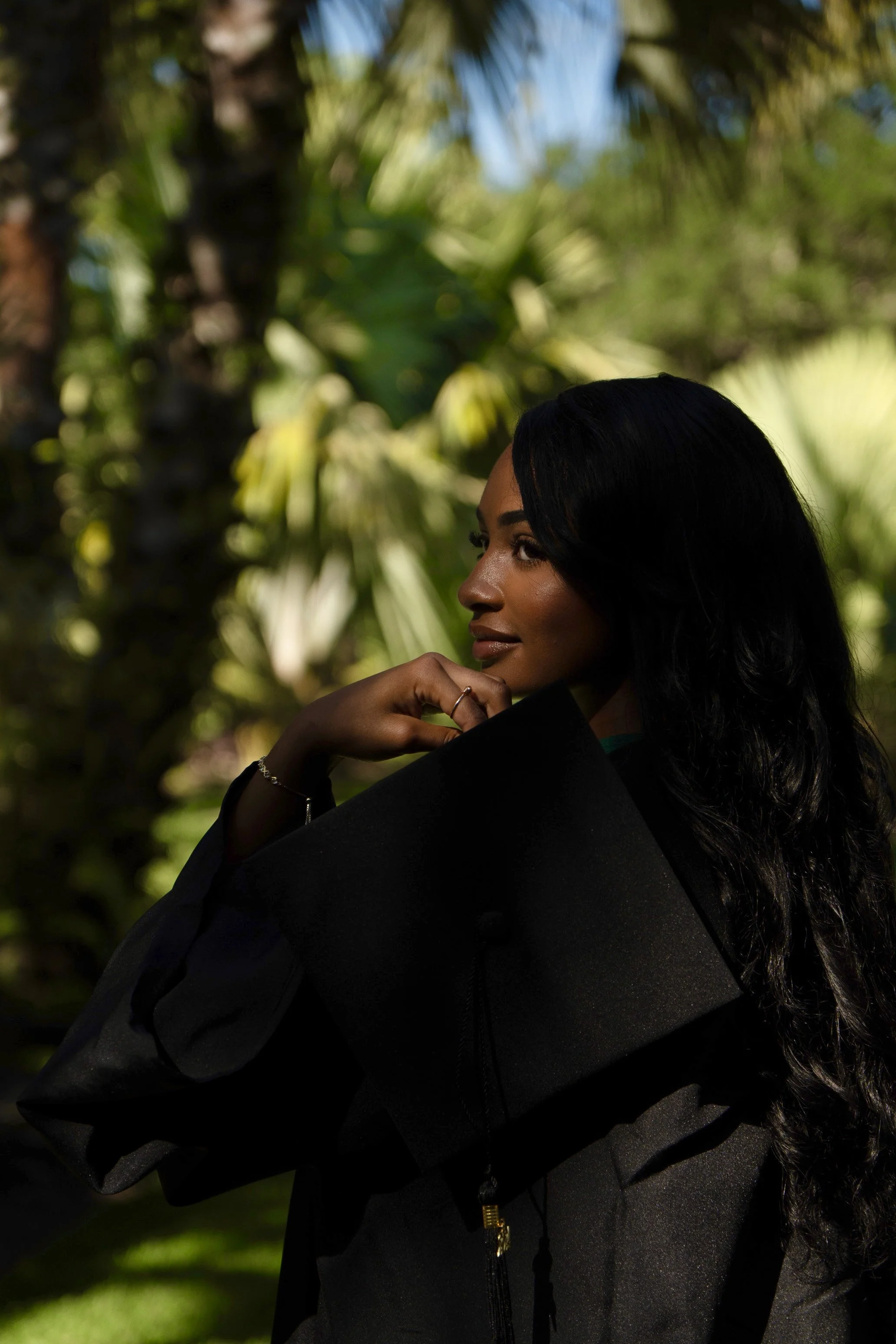 Graduate woman in black cap and gown outdoors with greenery backdrop.