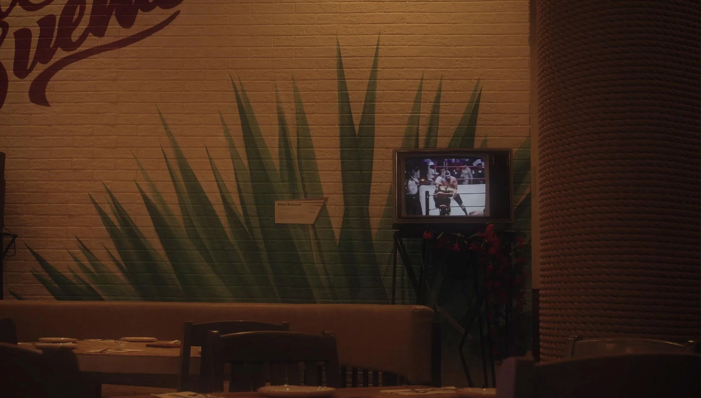 Dimly lit restaurant interior with agave mural, small TV showing wrestling match, wooden dining set in foreground.