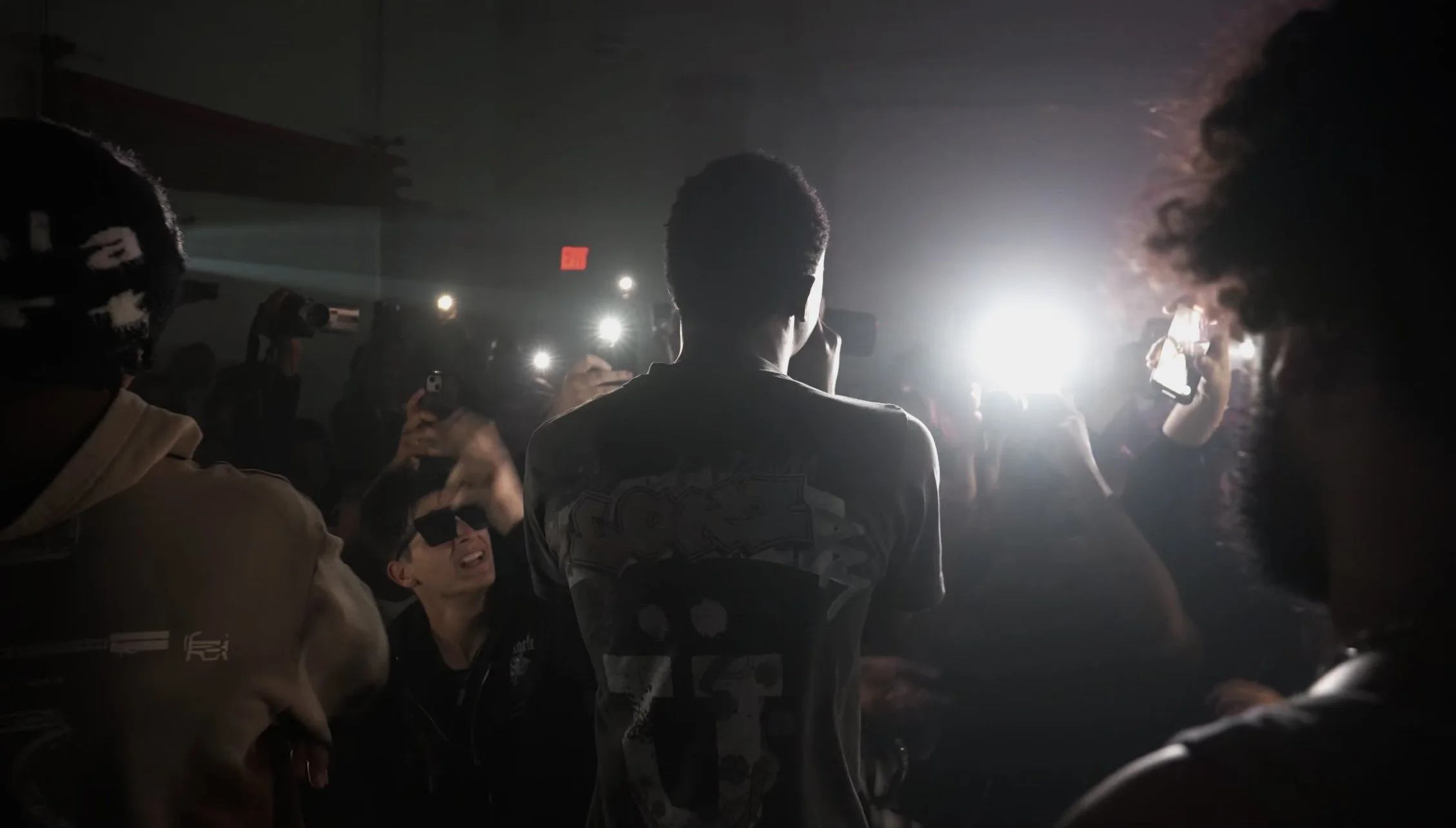 A back view of a performer on stage, with bright lights and a crowd taking photos with phones.