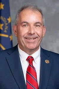 A man in a dark suit, white shirt, and red striped tie smiling in front of a gray background with an American flag and a Blue flag behind him.