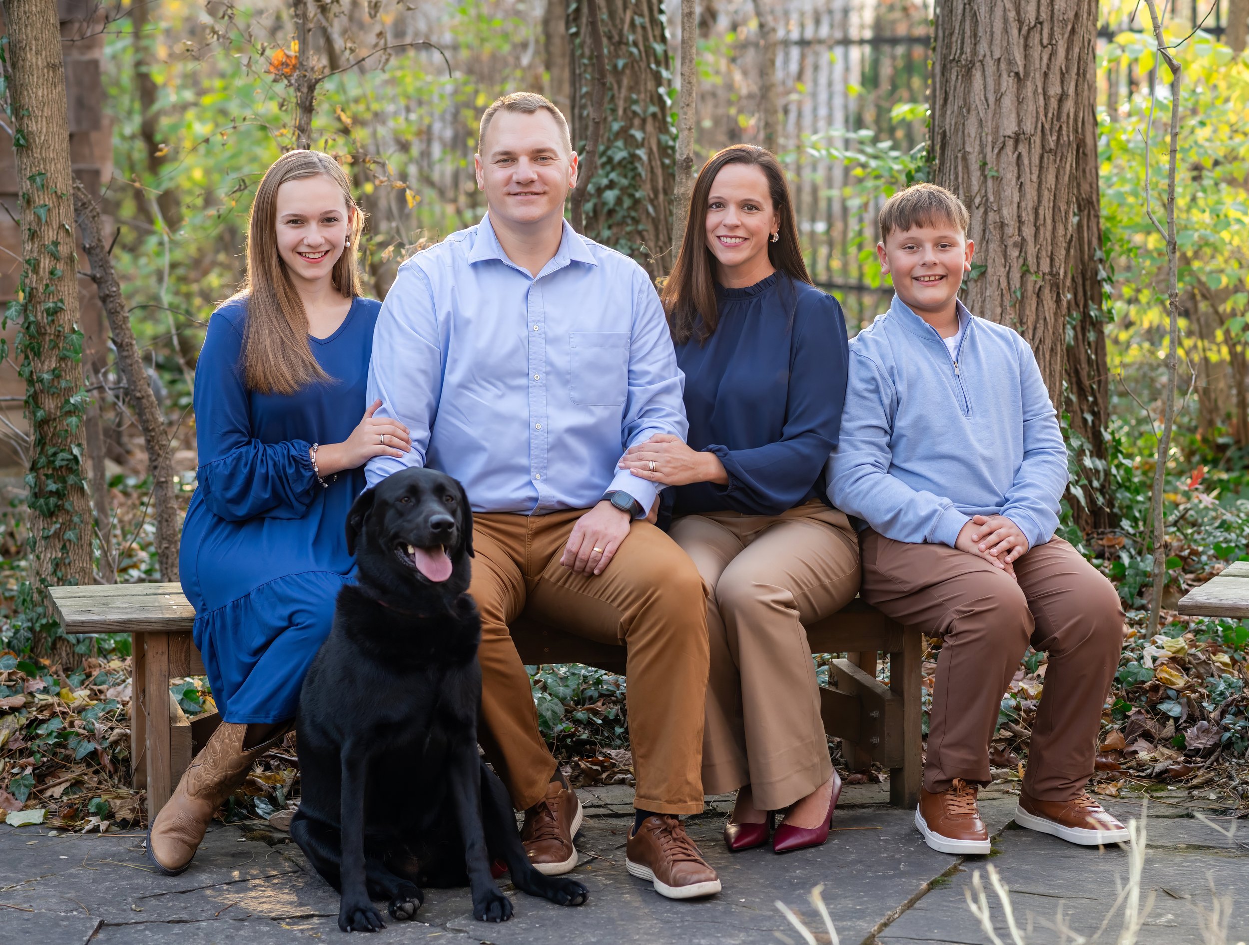 A family of four sitting on a wooden bench in a wooded outdoor area with a black dog in front. The family includes a girl, a man, a woman, and a boy all smiling at the camera.