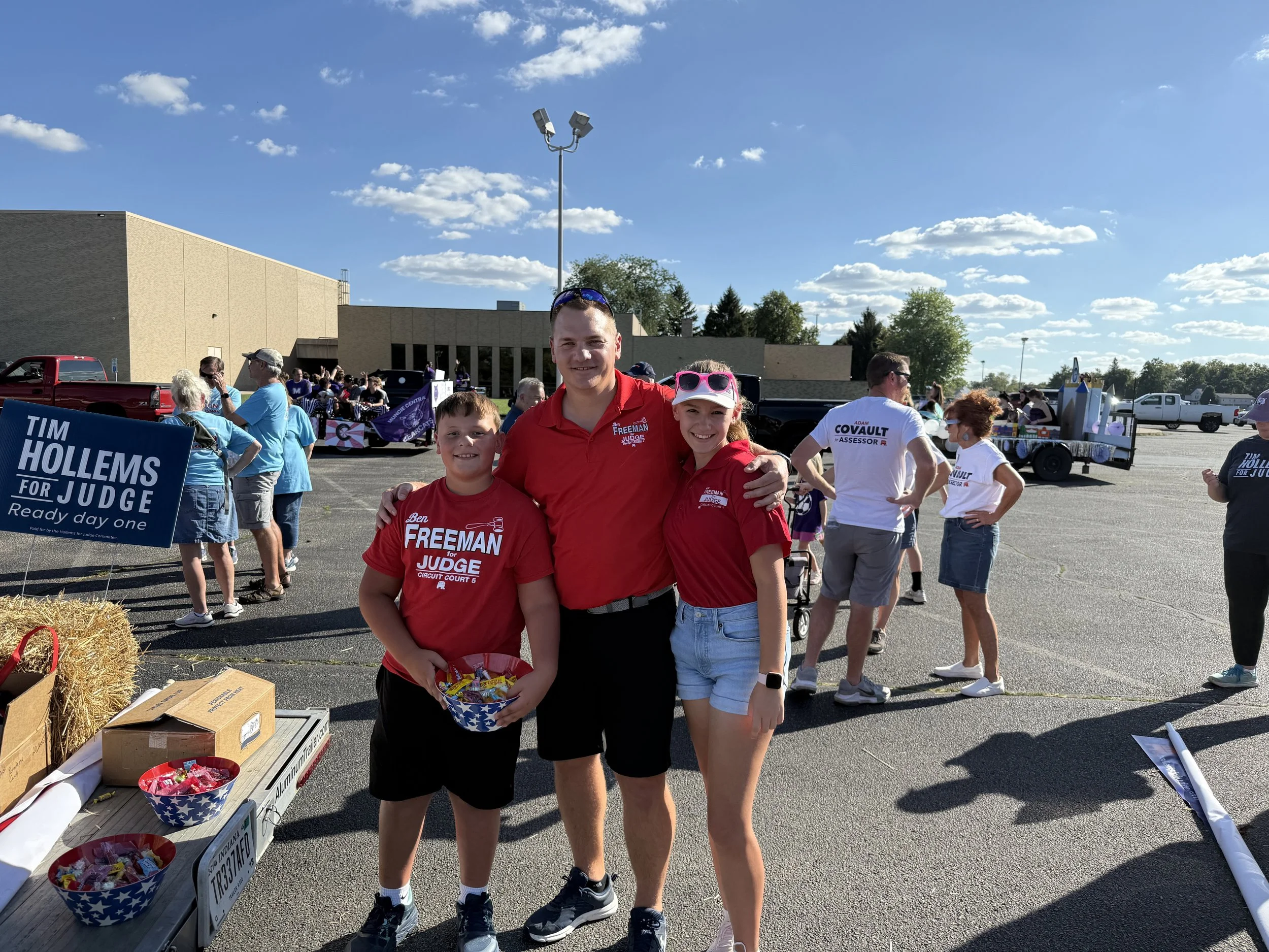 A group of three people, two boys and a woman, standing in a parking lot at an outdoor event. They are smiling, wearing red and in front of other event attendees and floats. The boy on the left is holding a bowl of candy, and the woman and the other 