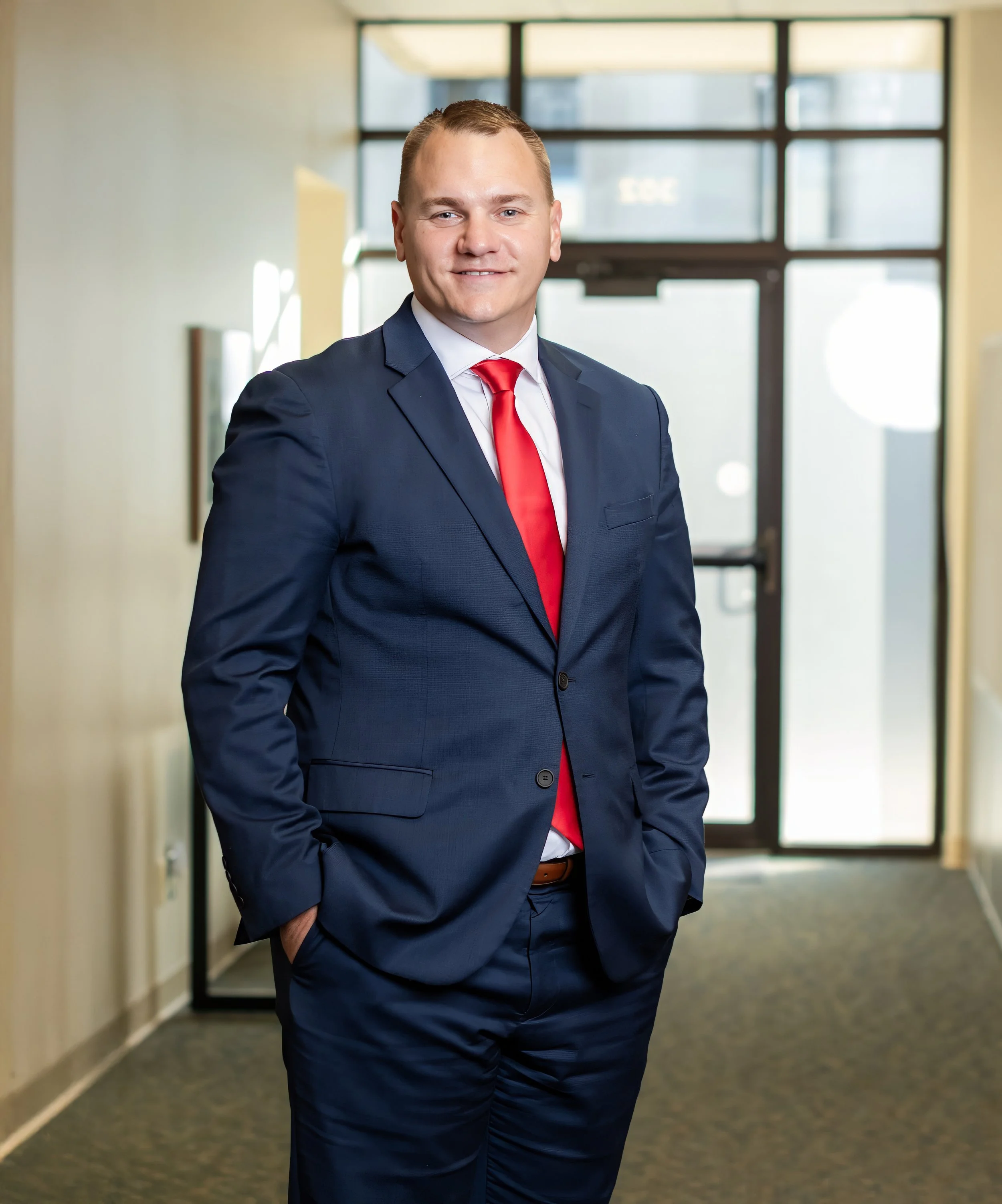 A man in a navy blue suit with a red tie and white shirt standing in a hallway with large windows behind him.