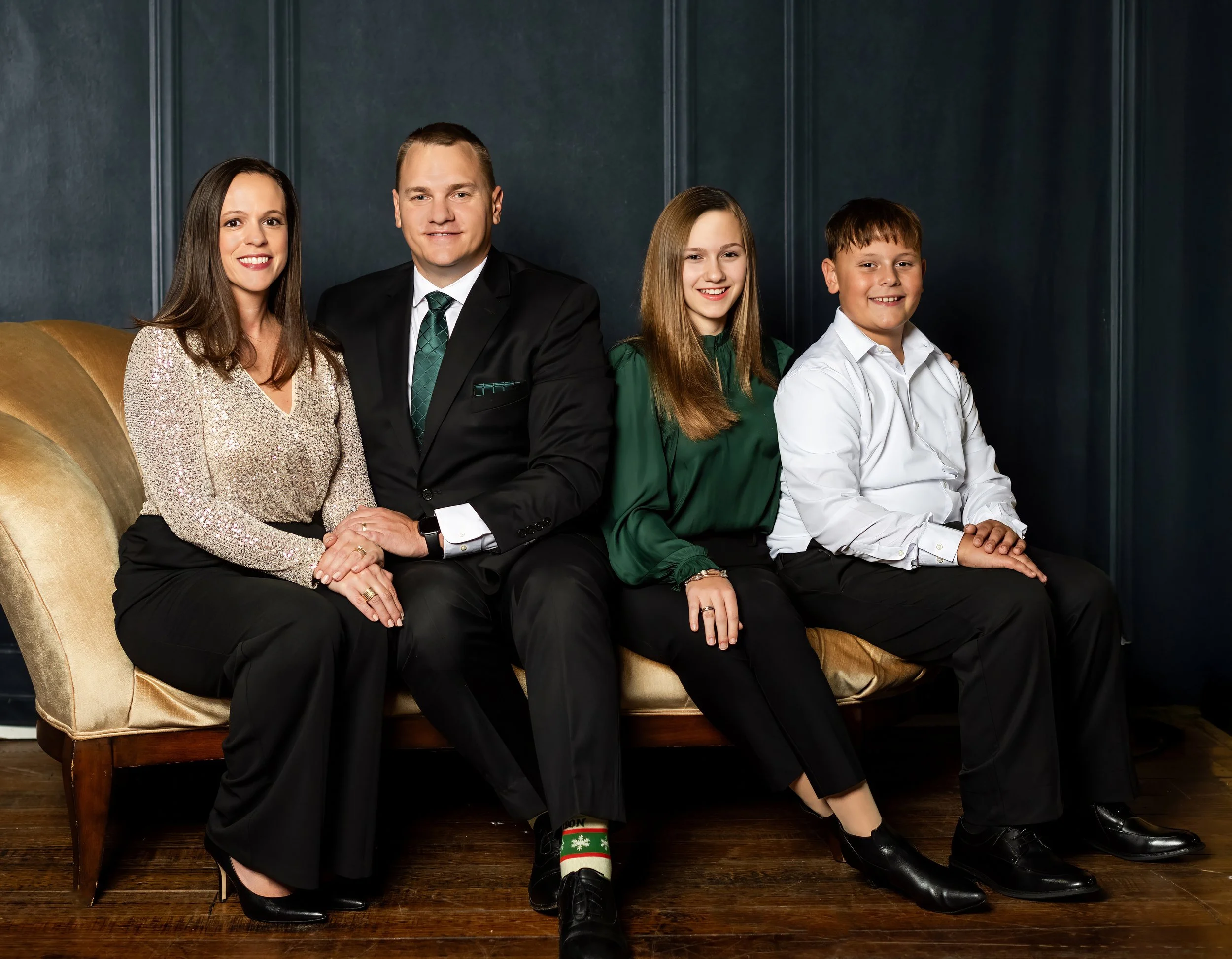 A family of four sitting on a vintage sofa in a dark green room, smiling for the camera. The mother is wearing a sparkly, light-colored blouse and black pants. The father is dressed in a black suit with a green tie. The daughter is wearing a green bl
