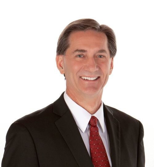 A smiling man in a black suit, white shirt, and red patterned tie with short, brown hair, against a white background.