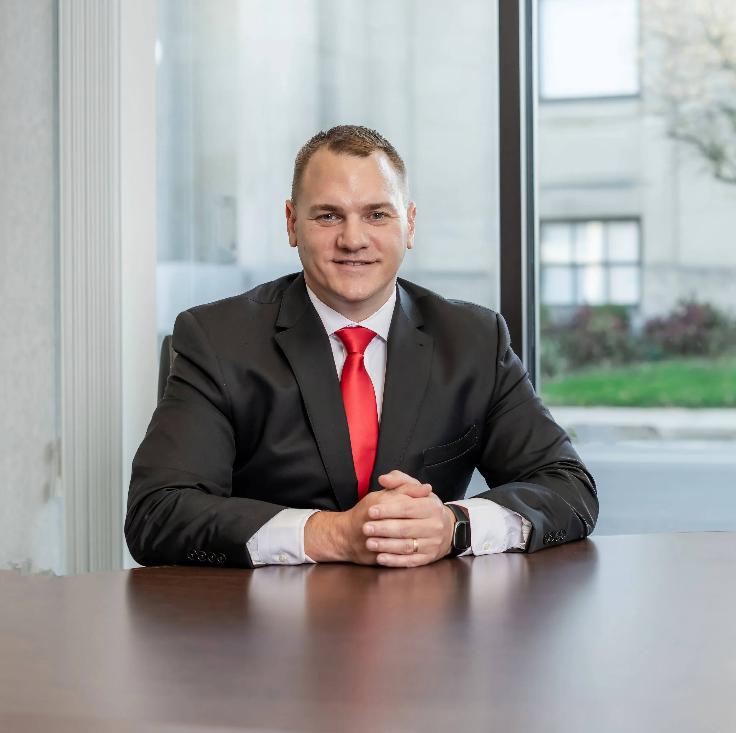 A man in a black suit, white shirt, and red tie sitting at a table in front of a window, smiling with hands clasped.
