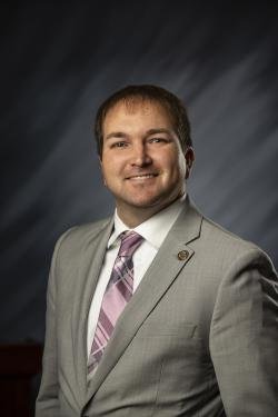 Portrait of a man in a light gray suit with a pink striped tie, smiling, against a dark, gradient background.