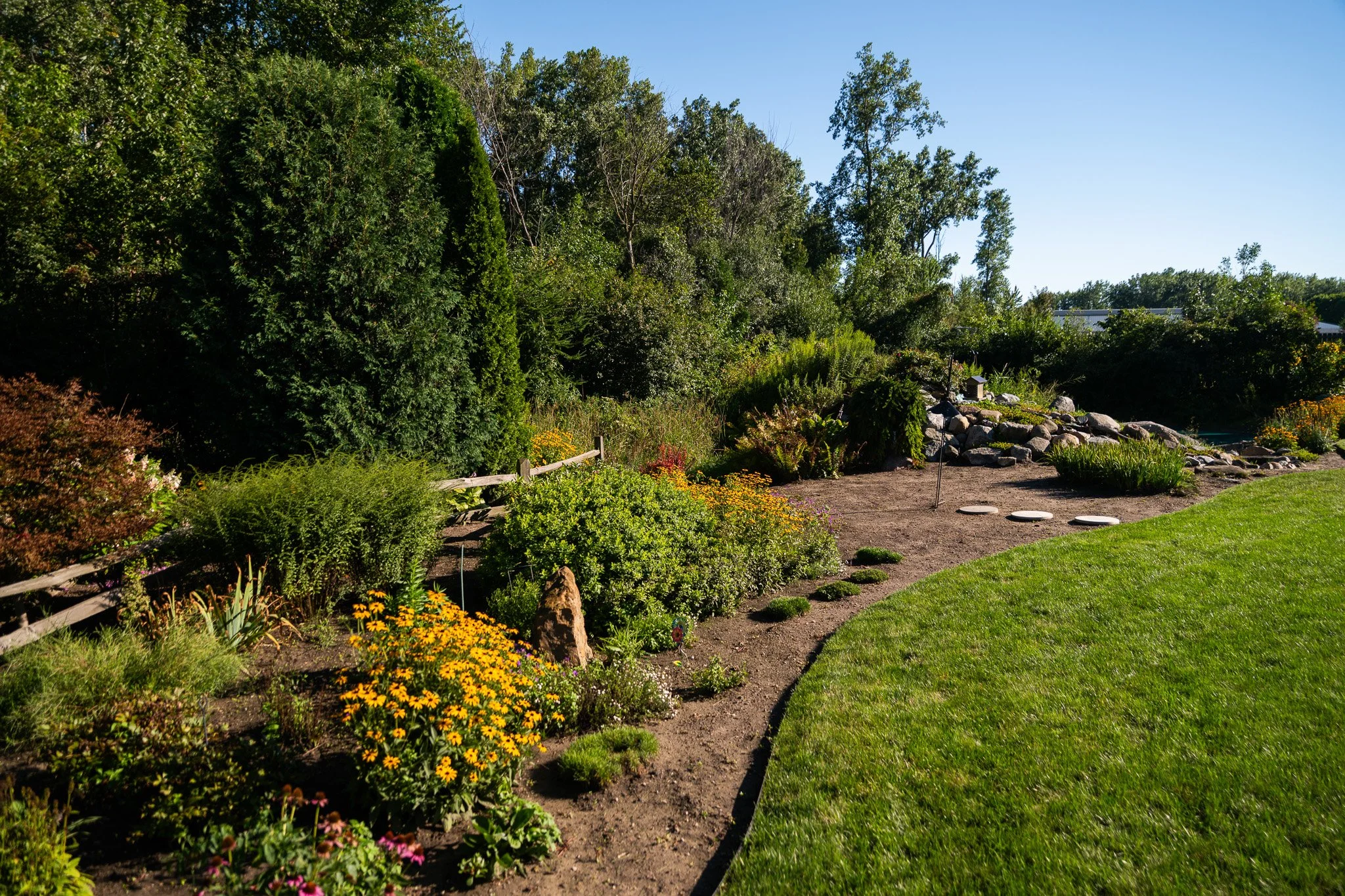 New Chapter Dentistry's landscaped garden with a dirt pathway surrounded by various green bushes, trees, and colorful flowers, with a grassy lawn on one side and rocks in the distance under a clear blue sky.