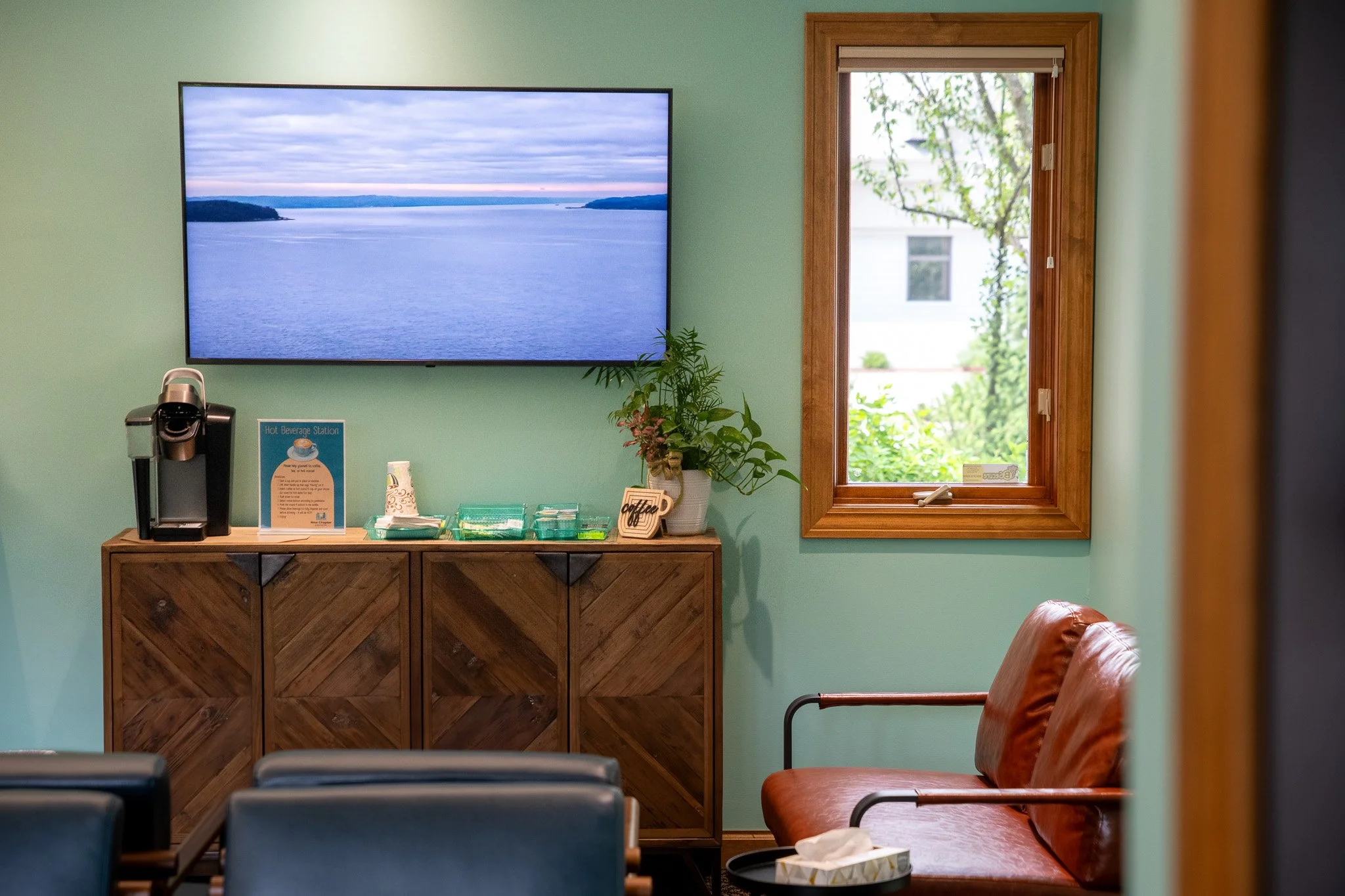 New Chapter Dentistry's receptionroom with a wall-mounted TV displaying a body of water and sky, a wooden cabinet with a coffee station and plant, a window with greenery outside, and two brown leather chairs.