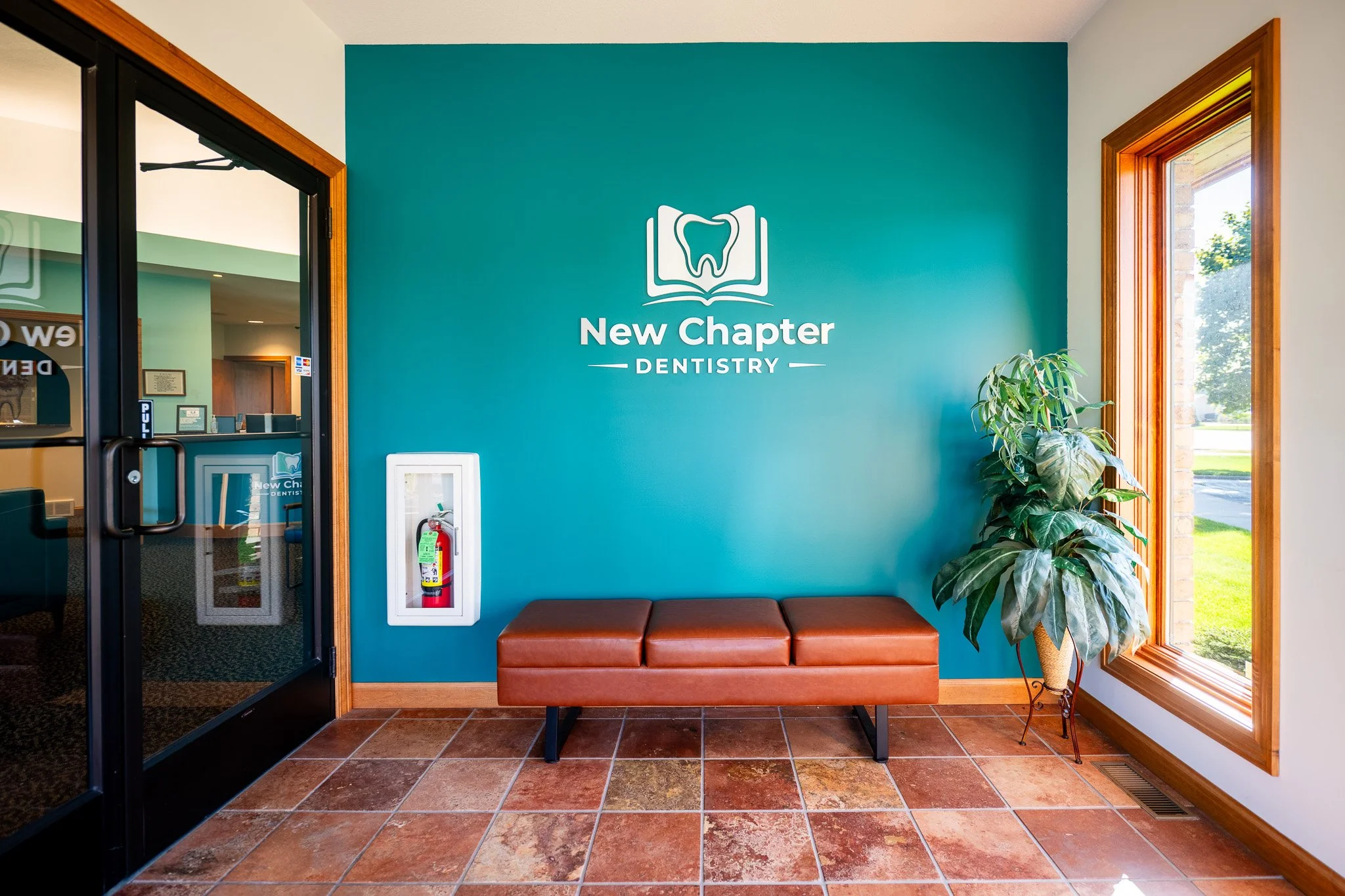 Interior of our dental office with a teal accent wall featuring a logo of a tooth reading 'New Chapter Dentistry', a brown bench, a potted plant, a window showing outdoor greenery, and a fire extinguisher.