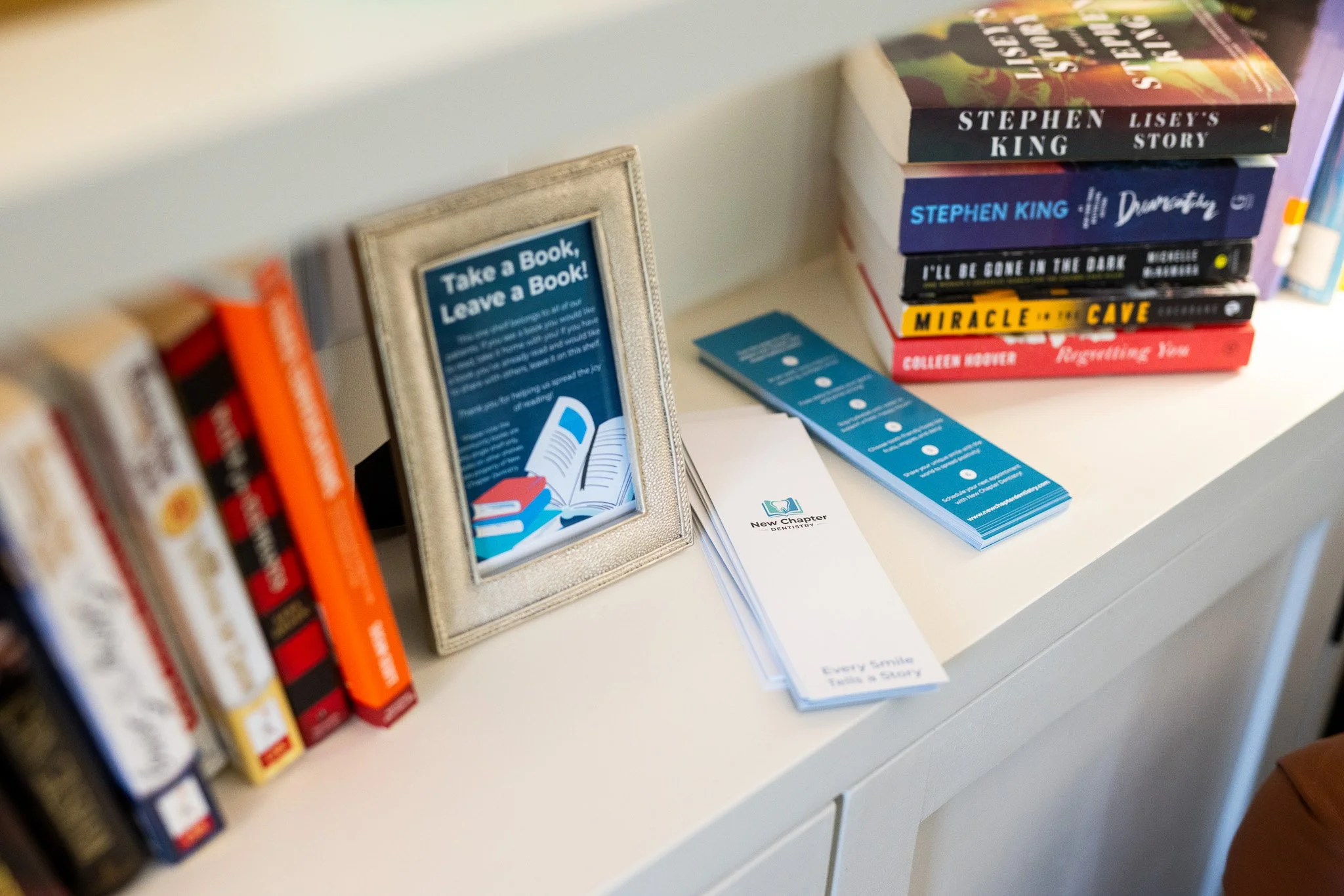 New Chapter Dentistry's library with a framed sign that says 'Take a Book, Leave a Book!' and a stack of books, along with pamphlets and informational flyers.