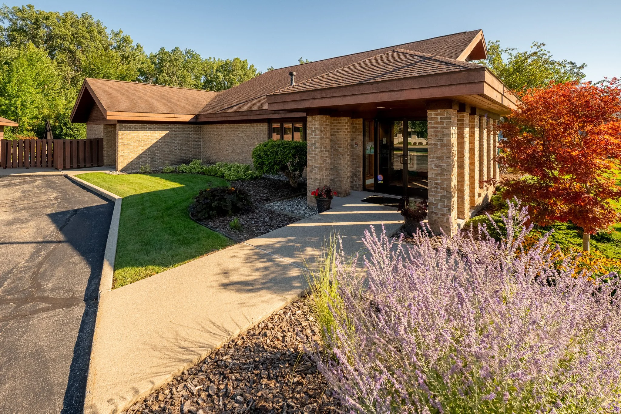 A brick building with a brown shingled roof and glass front door. The landscaped front yard has green grass, bushes, purple flowers, and a tree with red leaves. A paved driveway and sidewalk lead to the entrance.