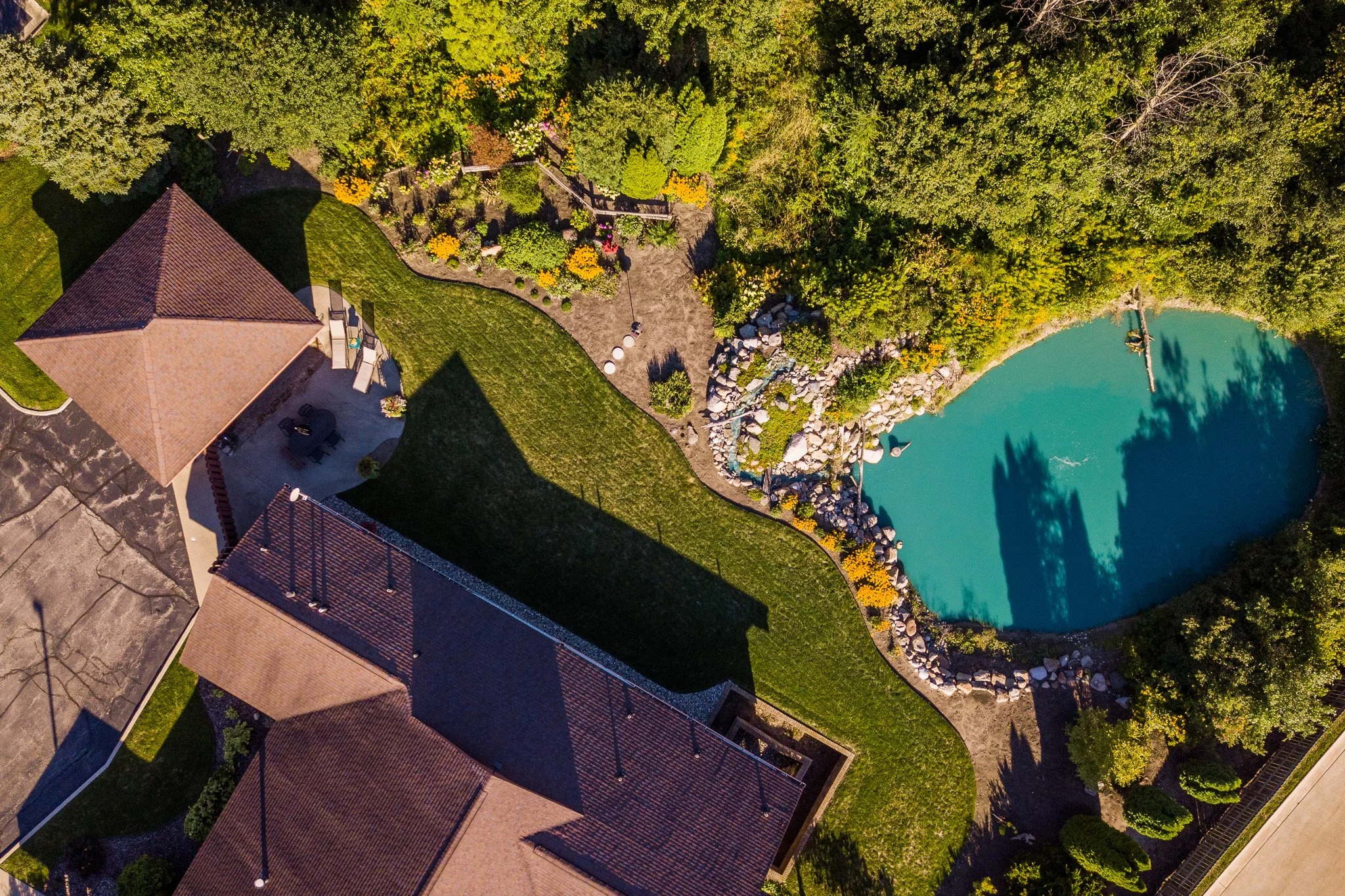 An aerial view of a backyard with a swimming pool, green grass, a patio with outdoor furniture, a garden with colorful flowers, and a wooded area surrounding the yard.
