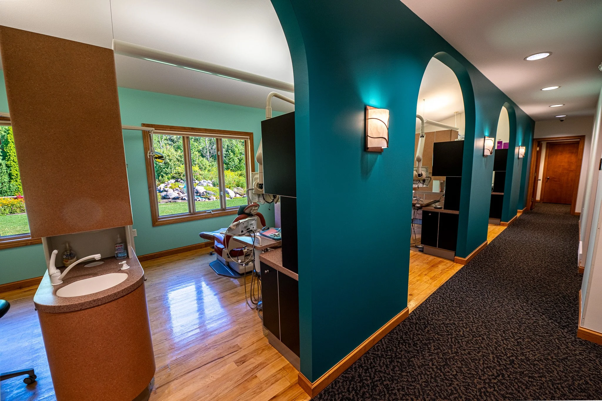 New Chapter Dentistry's treatment room with dental chair, medical equipment, and large windows overlooking a garden, divided by teal walls with wall sconces.