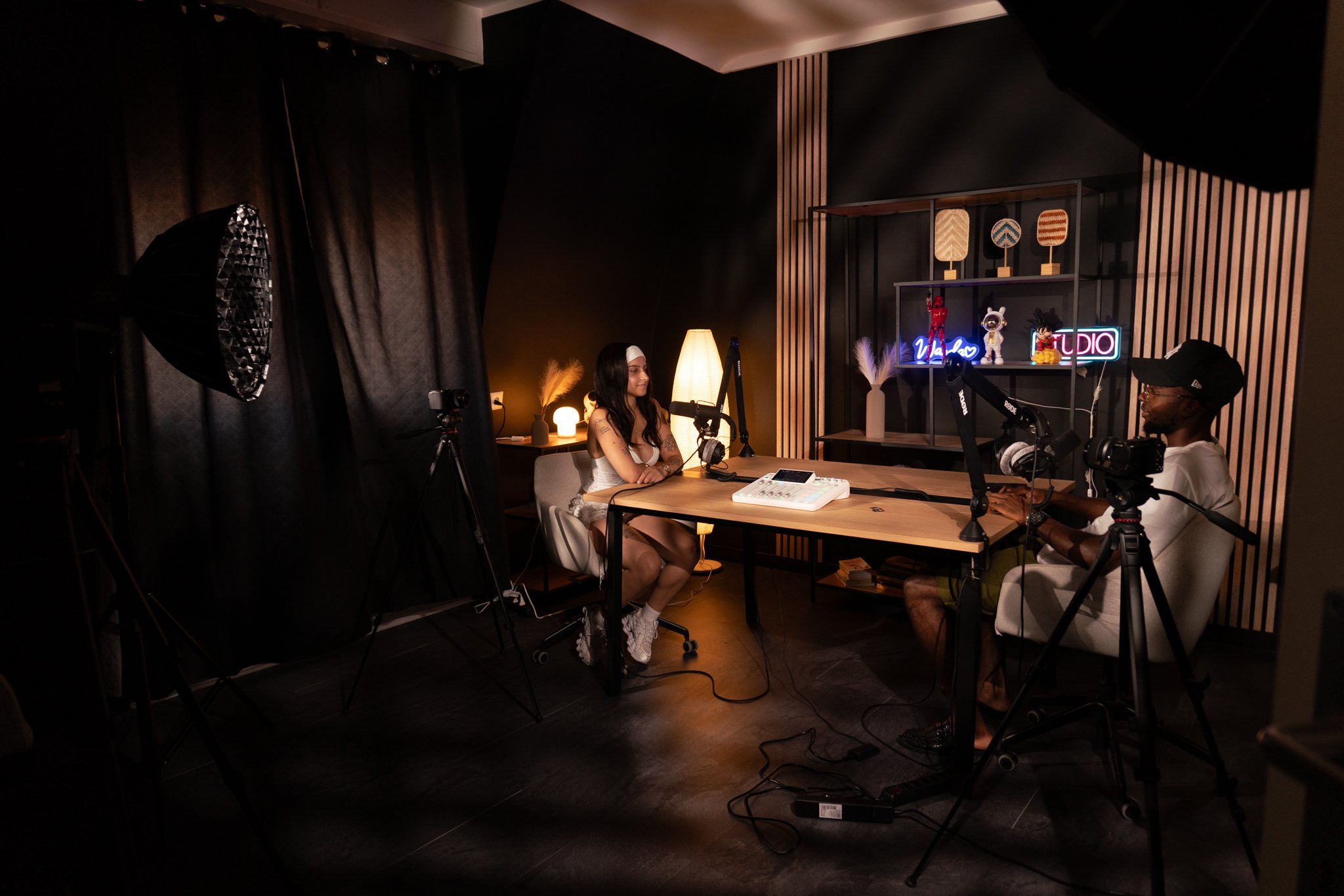 Une femme et un homme enregistrent une émission ou un podcast dans un studio avec des équipements de tournage, des microphones, des caméras et un décor moderne et chaleureux.