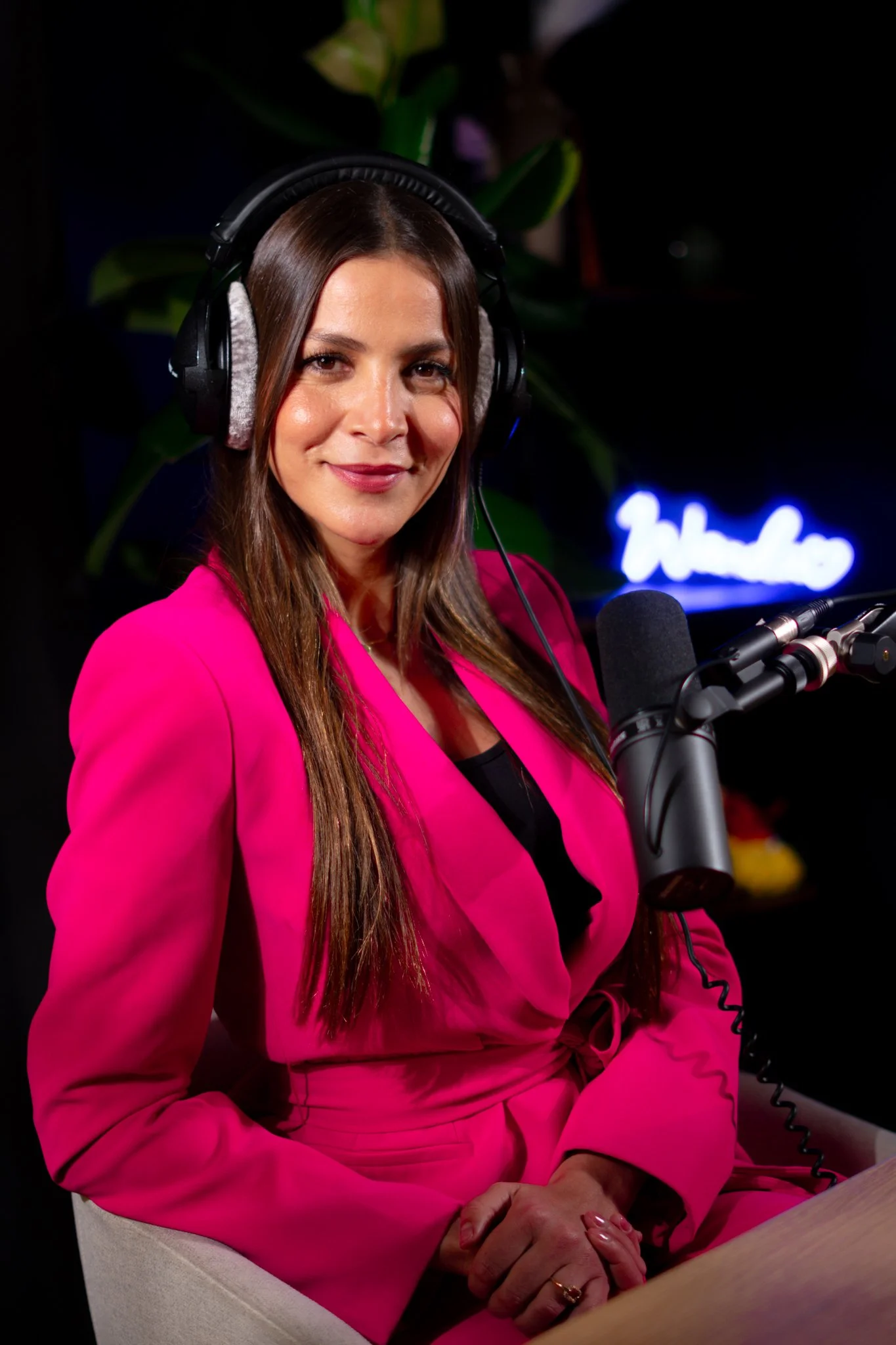 Femme souriante portant un costume rose et un casque, assise devant un microphone dans un studio d'enregistrement