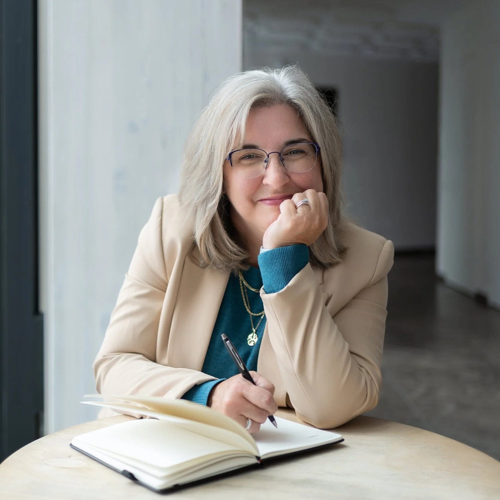 A smiling middle-aged woman with gray hair and glasses sitting at a table, holding a pen and writing in a notebook.