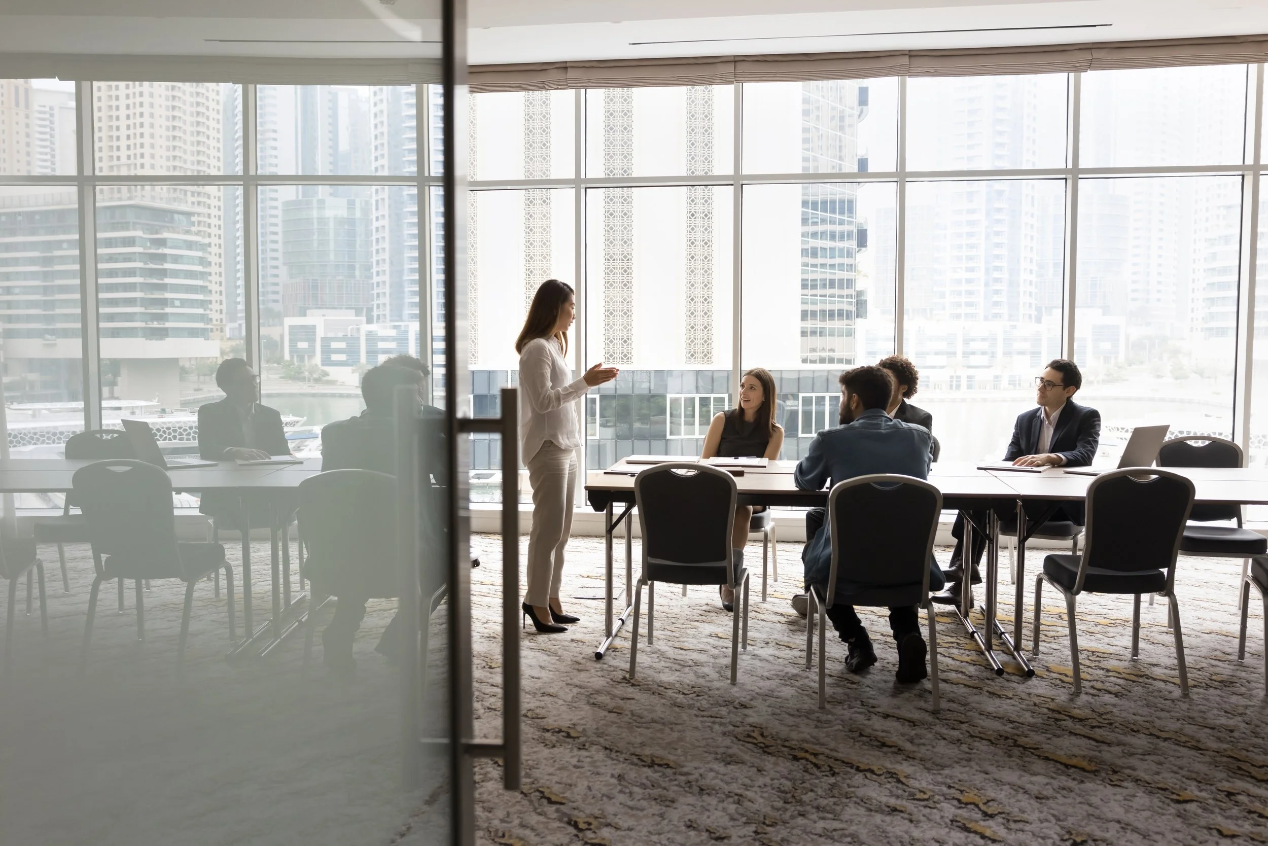A woman standing and speaking to a group of five people sitting at a conference table in a modern office with large windows overlooking a city skyline.