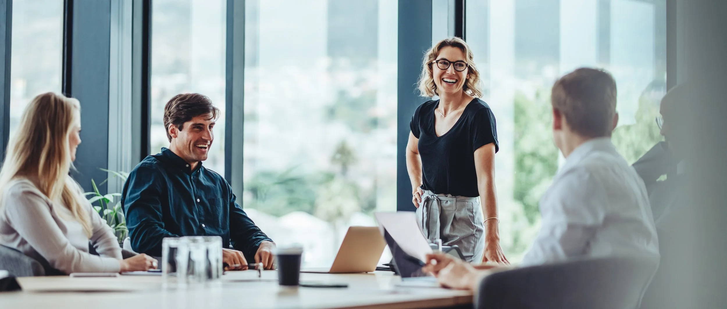 Group of professionals in a business meeting around a conference table, with a woman standing and speaking, all smiling.