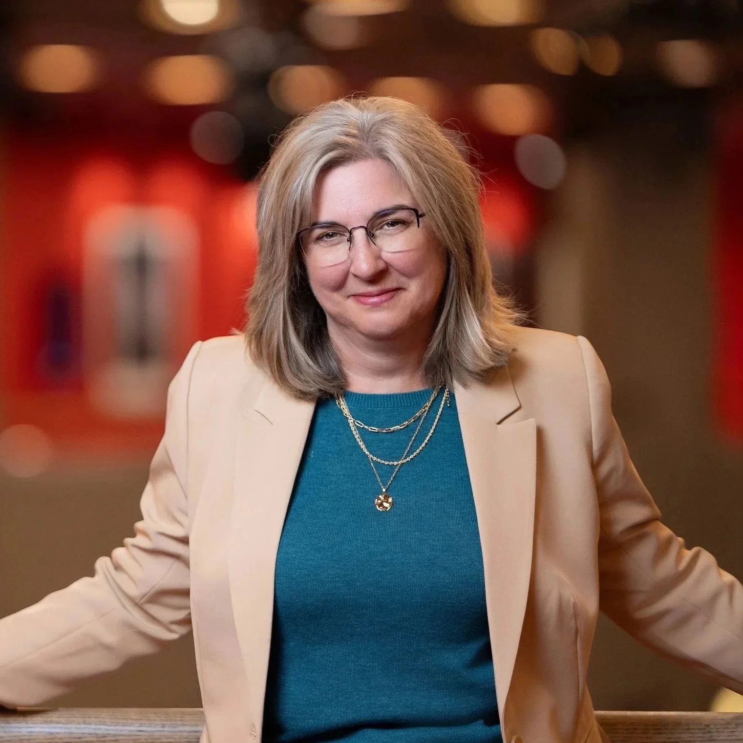 A woman with shoulder-length gray hair and glasses, wearing a beige blazer over a teal top and layered necklaces, standing with arms outstretched in an indoor setting with blurred colorful lights in the background.