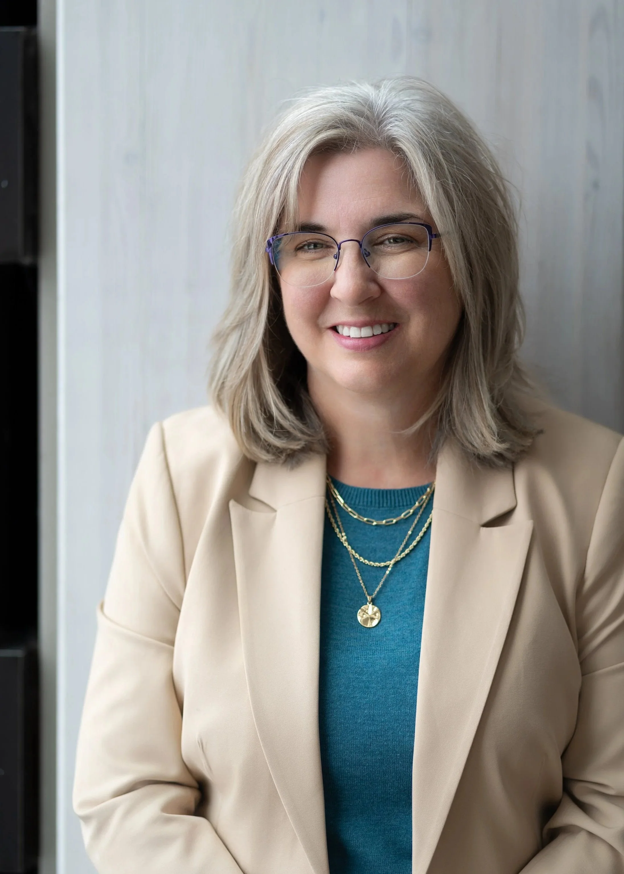 A woman with shoulder-length gray hair, wearing glasses, a beige blazer, and a blue shirt, smiling in front of a light-colored wall.