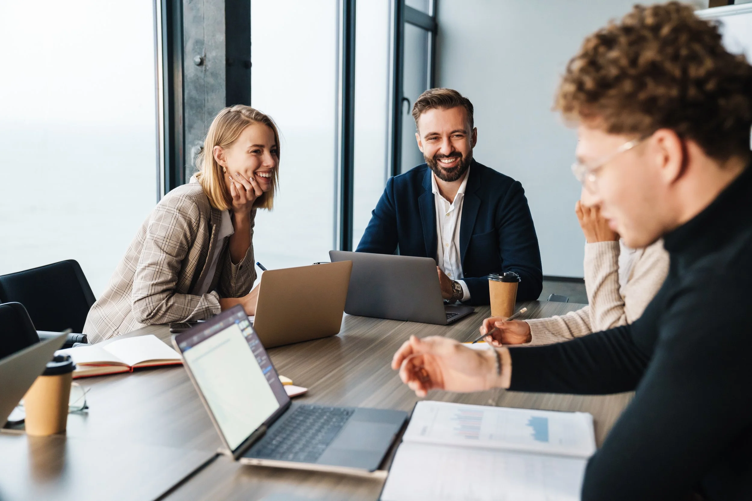 Four professionals sitting at a conference table in a modern office, smiling and engaging in a discussion, with laptops, notebooks, and coffee cups on the table.