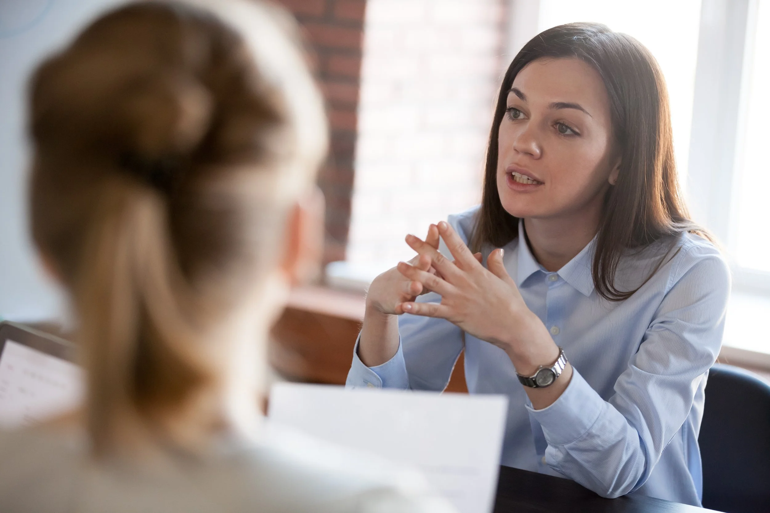 Two women having a conversation indoors, woman in focus on the right with dark hair, wearing a light blue button-up shirt, appears to be speaking, woman in the foreground with layered blonde hair, blurred.