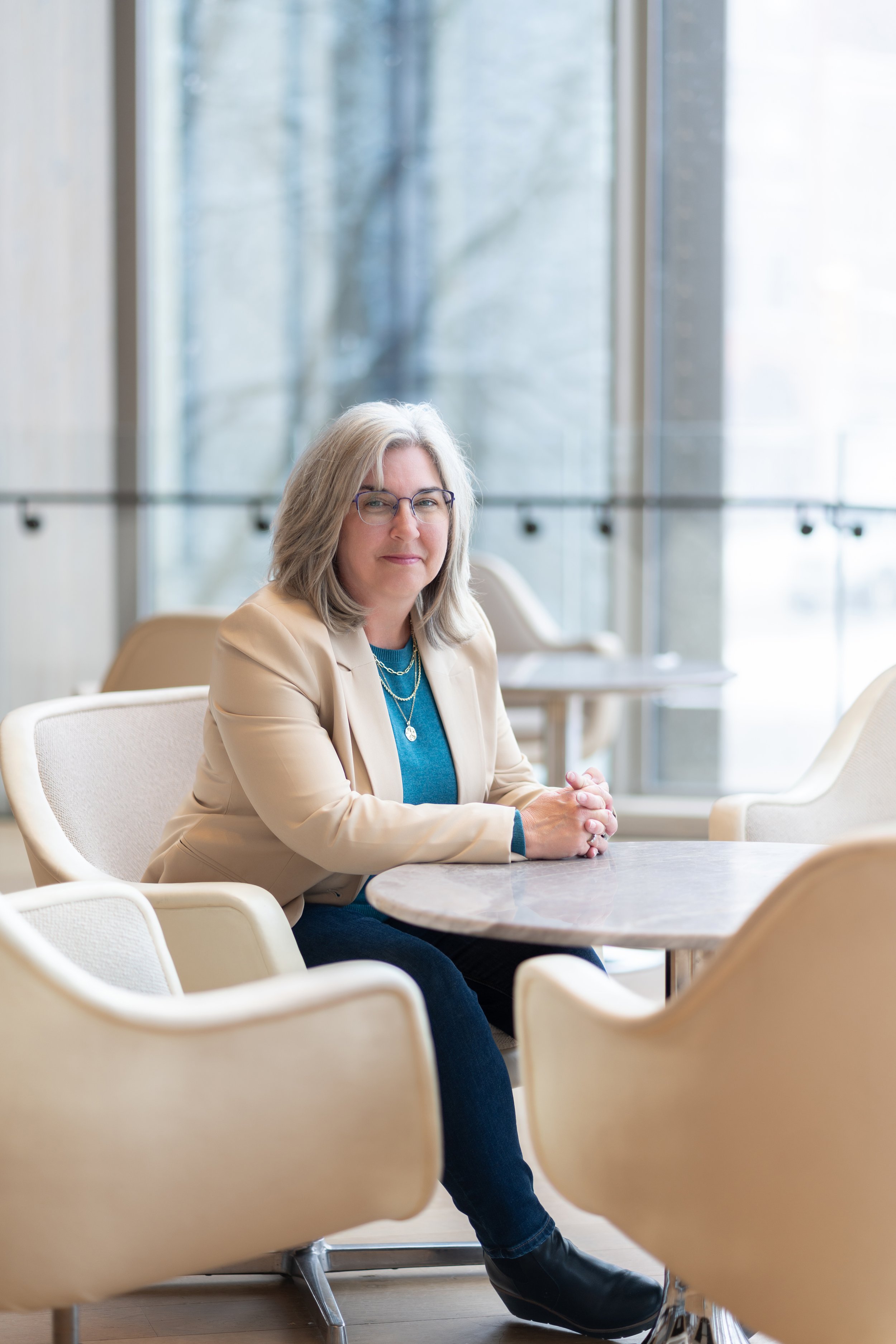Sharon McLaughlin sitting at a round table in a modern, well-lit cafe or lobby area with large windows and light-colored furniture.
