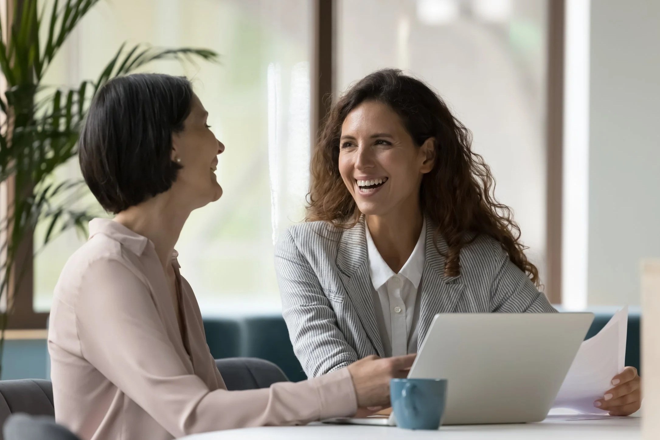 Two women sitting at a table in a bright office, smiling and talking while looking at each other. One woman has dark hair in a bob and the other has curly brown hair. There is a laptop and a blue mug on the table.