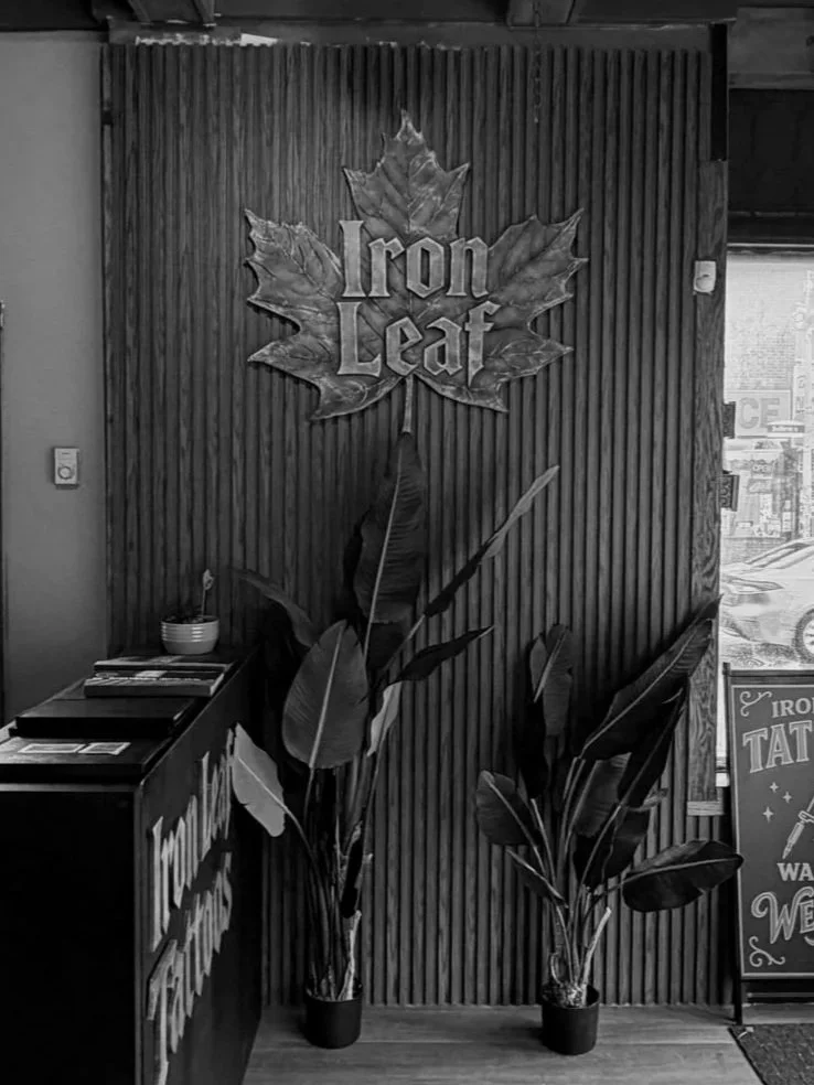 Black and white photo of a wall with a large Iron Leaf sign made of metal leaves, flanked by potted plants, with a reception desk on the left and a window to the right displaying street signs.