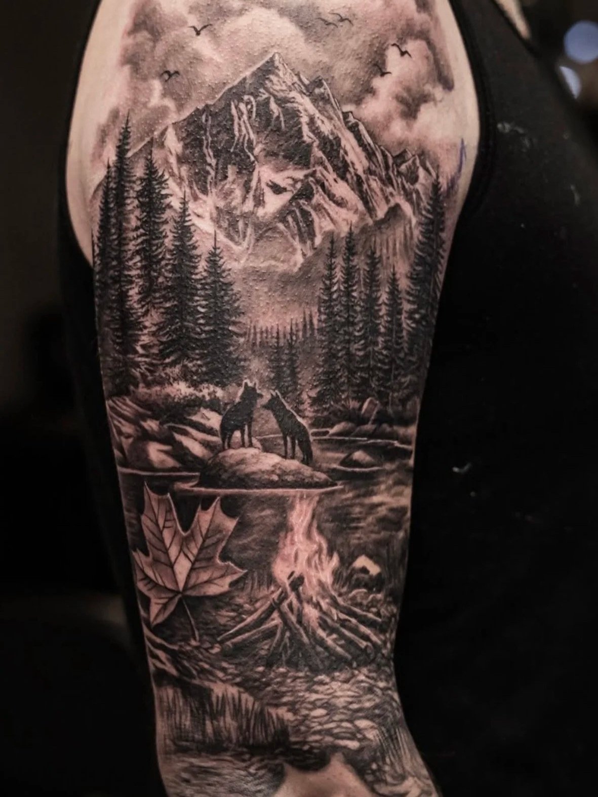 Two wolves on rocks with camp fire and maple leaf with trees and mountain background tattoo