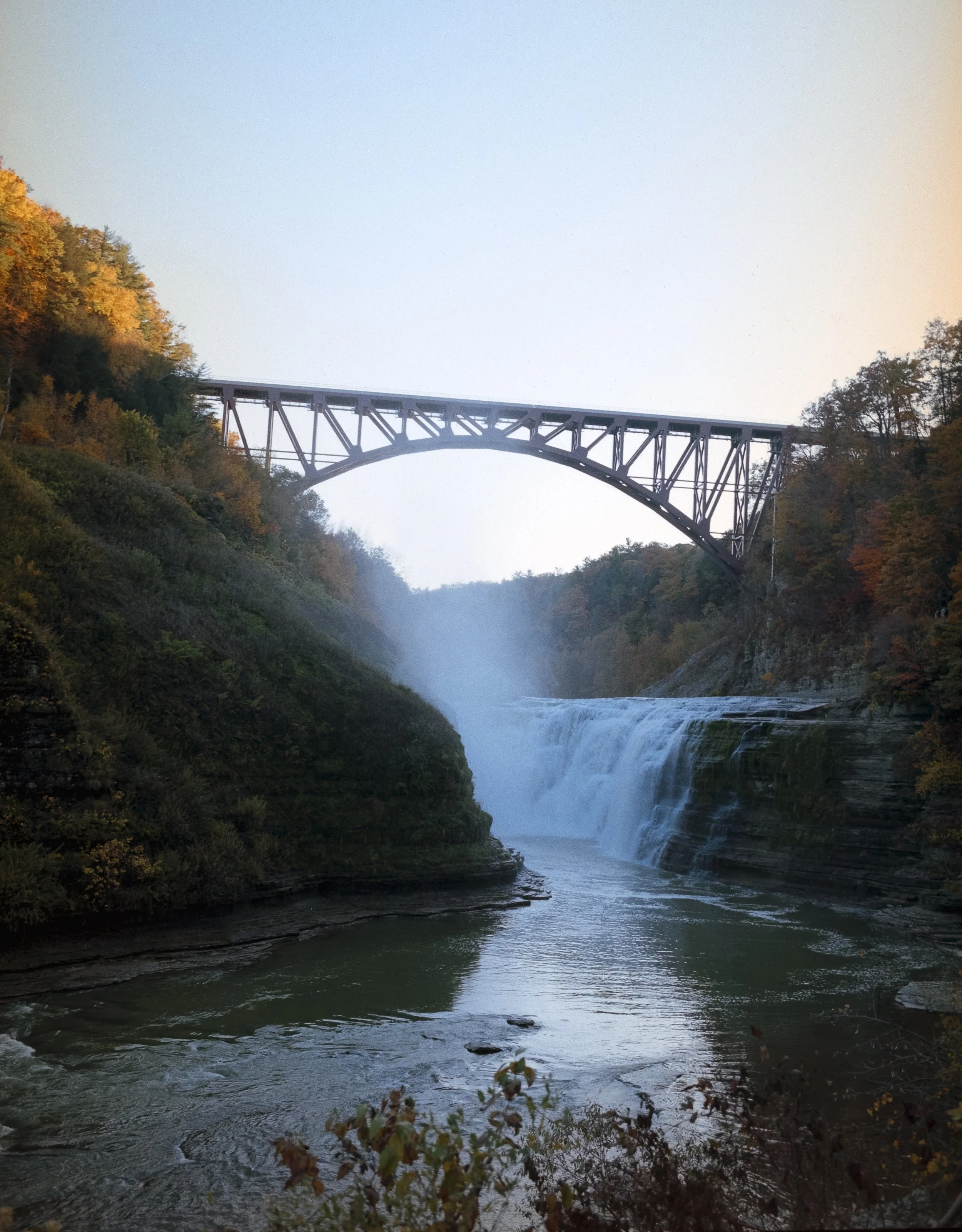 A scenic view of a waterfall with a bridge arching over it, surrounded by autumn-colored trees.