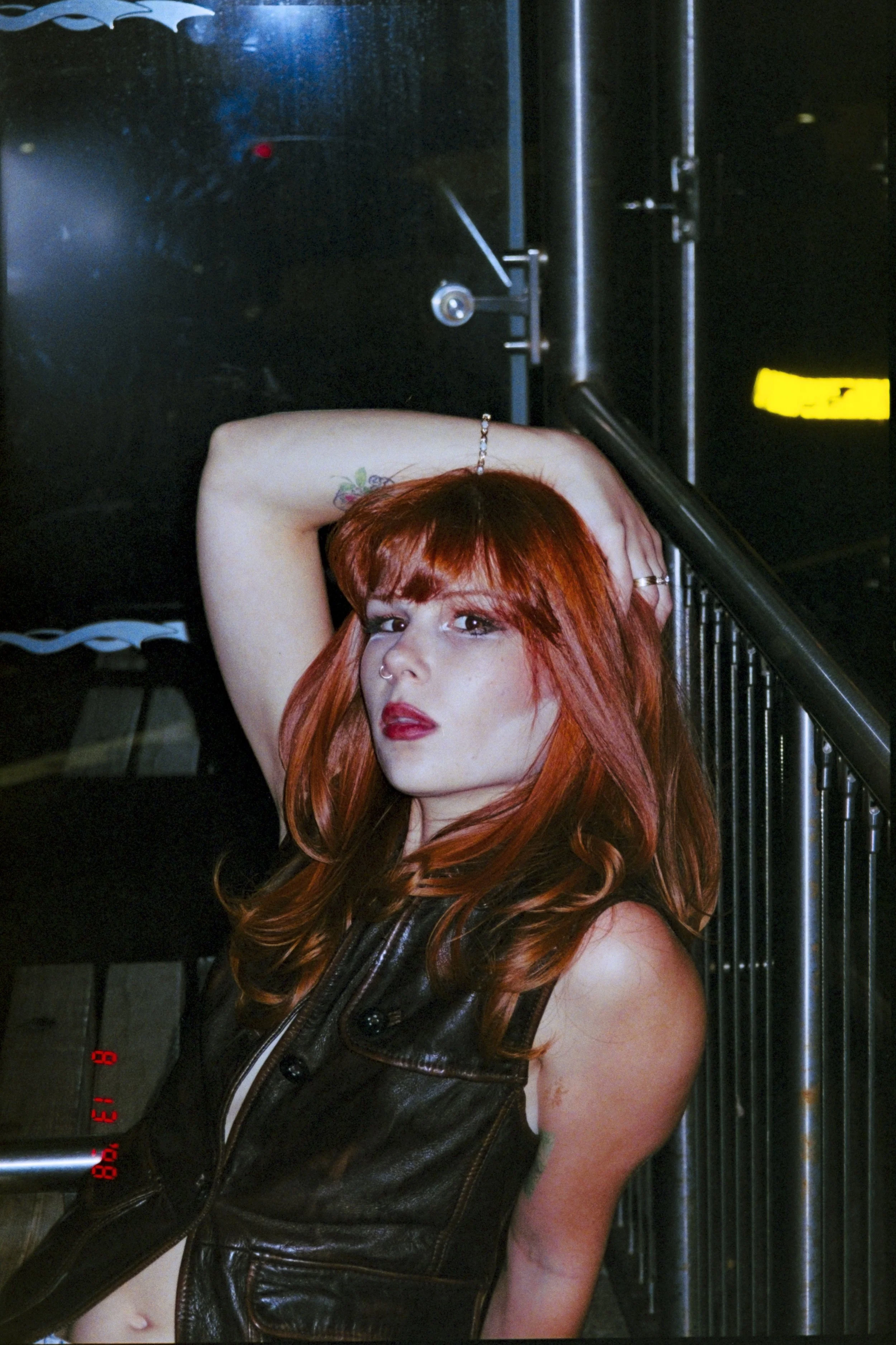 A young woman with long red hair and a septum piercing, standing next to a black metal railing, resting her head on her hand with her arm raised. She is wearing a black leather vest and has tattoos on her arm. The background shows a dark, reflective 