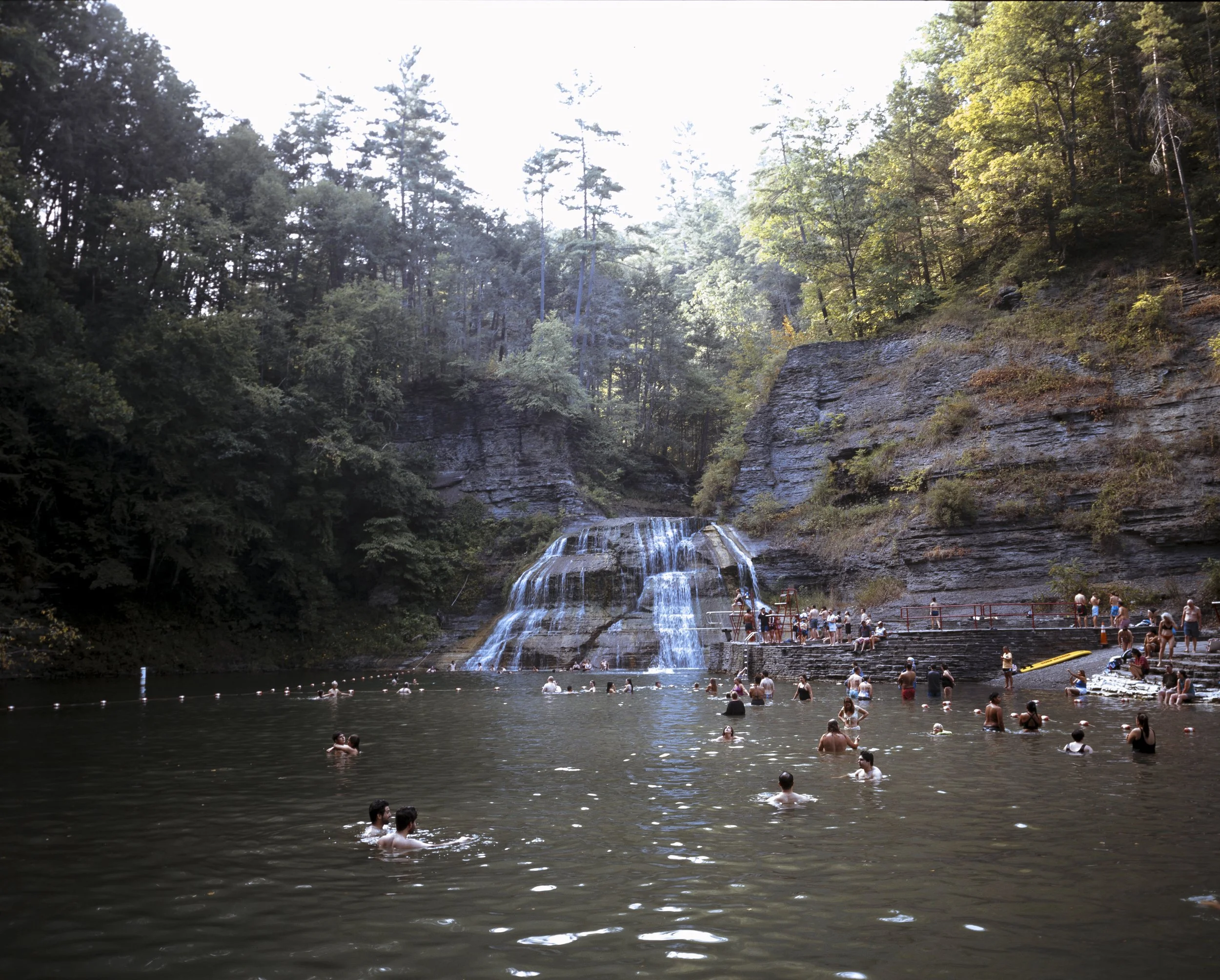 A popular swimming hole with people swimming and relaxing, surrounded by dense forest and a rocky waterfall creating a scenic natural setting.