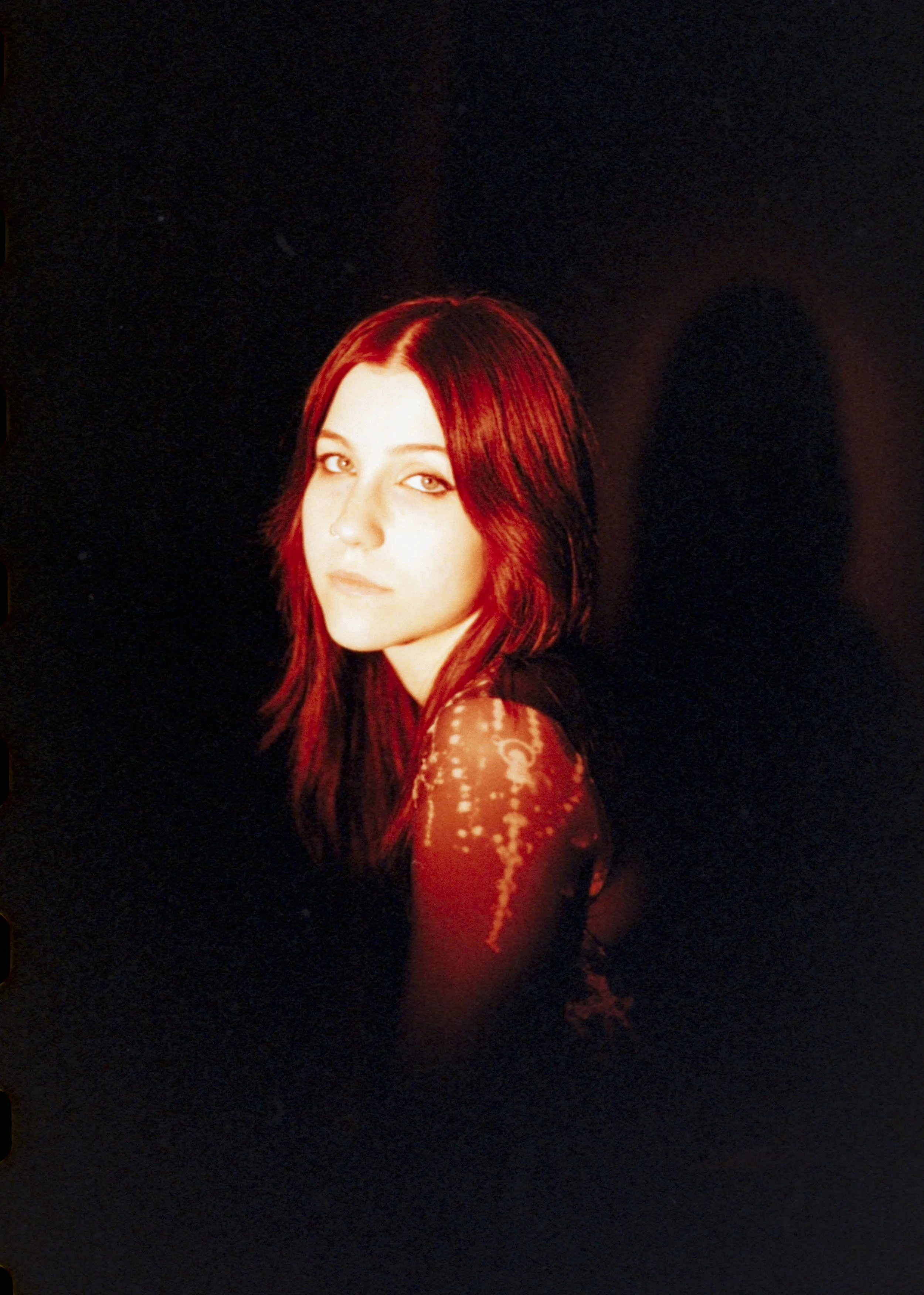 Portrait of a woman with reddish-brown hair and a serious expression, wearing a dark top with a shimmering pattern on the sleeve, against a black background.
