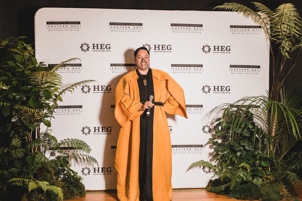 A man in a yellow gown standing on stage with a microphone, smiling, in front of a white backdrop with logos for NZ Chambers of Commerce Eastern Bay and Horizon Energy Group. There are green plants on either side of him.