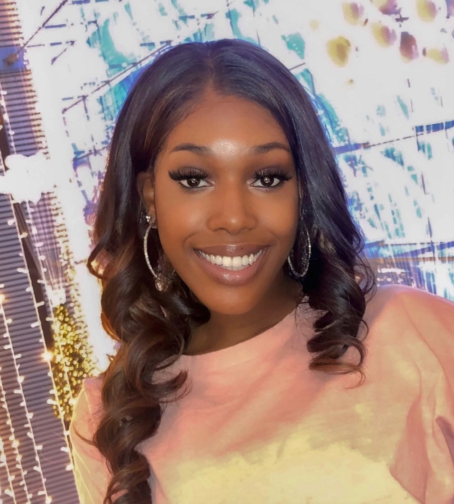 A young woman with long dark curly hair, hoop earrings, and makeup, smiling at the camera with a colorful abstract background.
