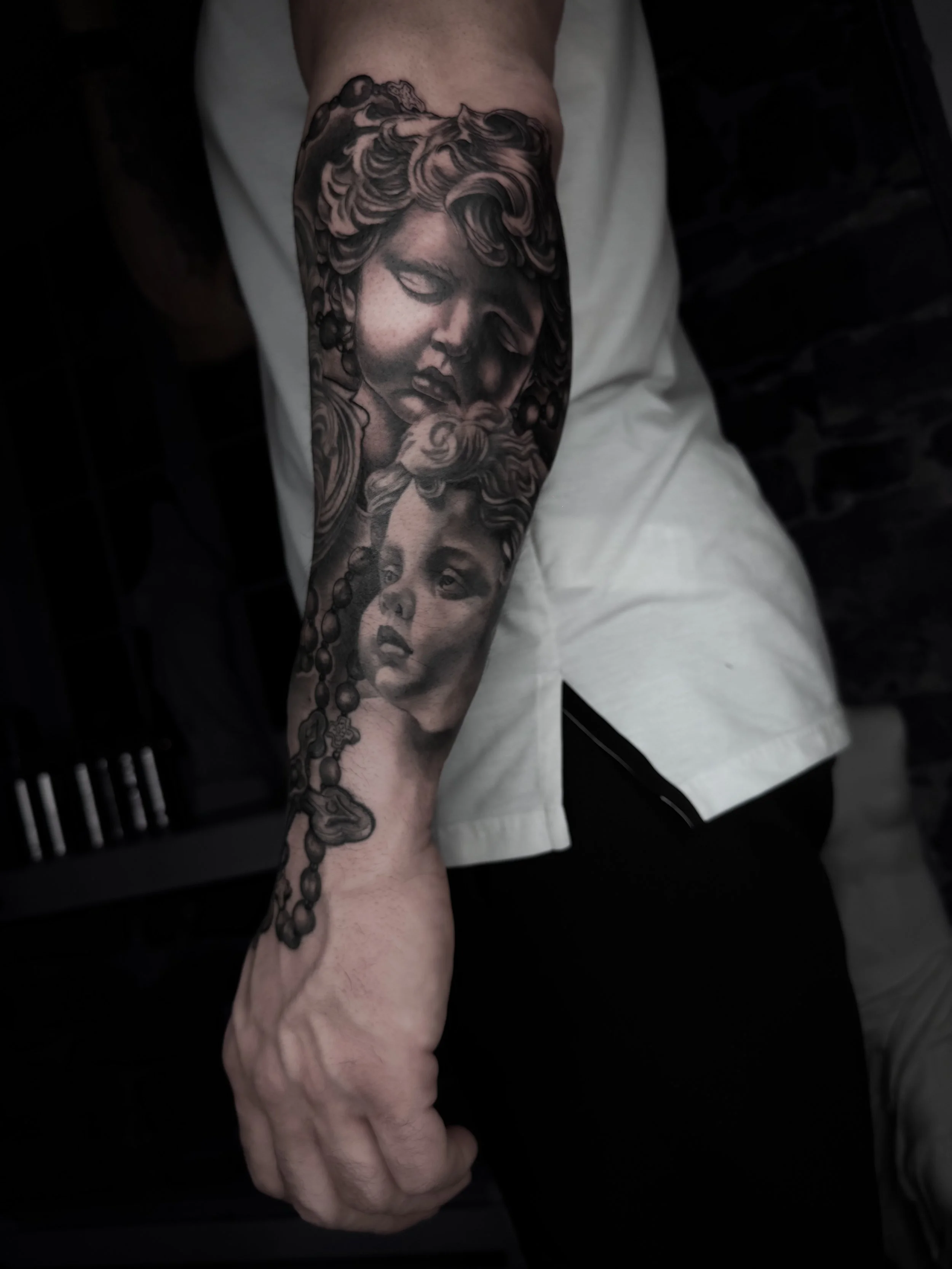 Tatouage avant-bras homme noir et gris réaliste – deux chérubins et chapelet, style religieux baroque. Darick Tattoos, montreal