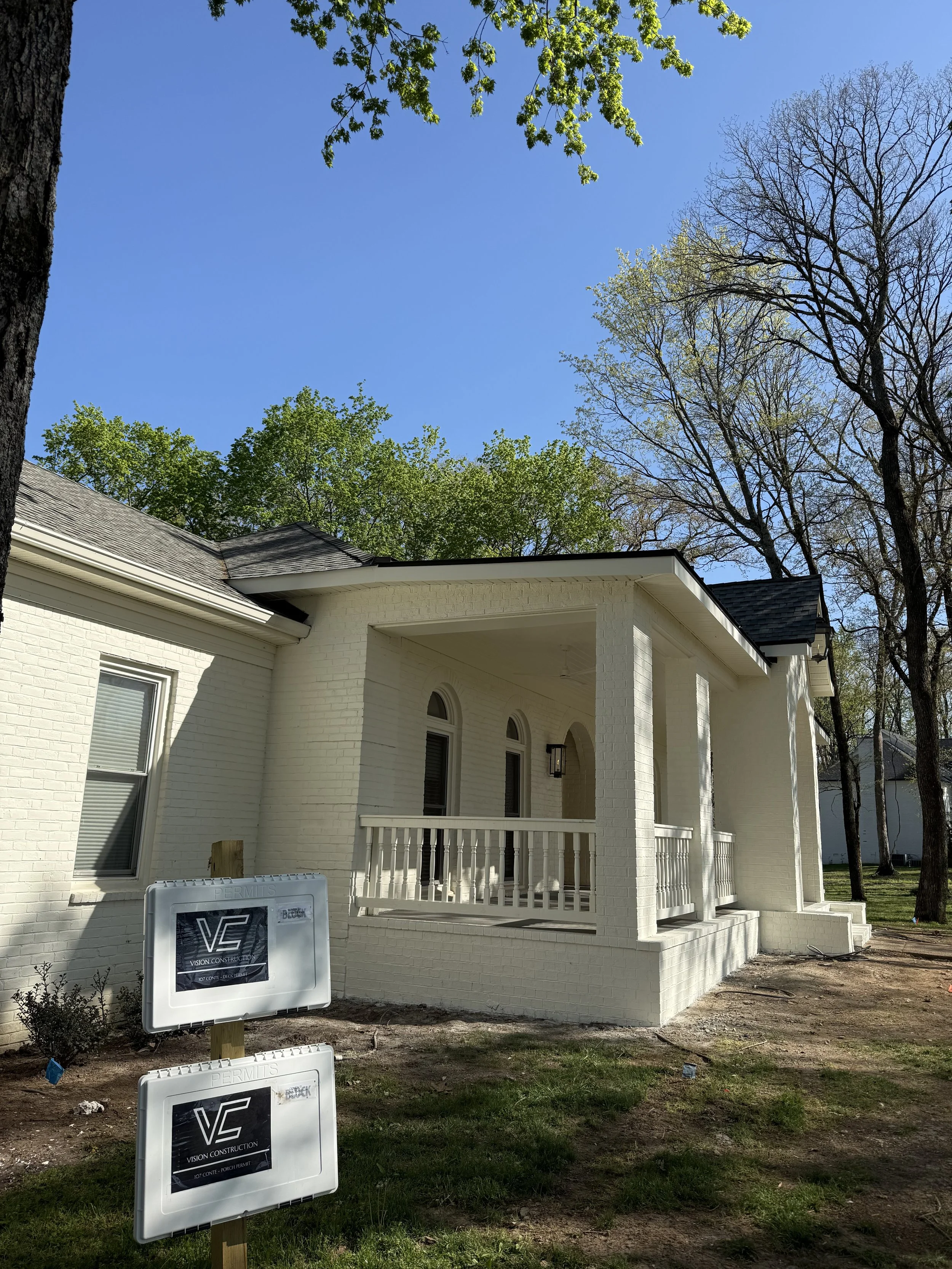Newly built white brick house with a front porch, surrounded by green trees, on a clear day. Construction signs are in the front yard.