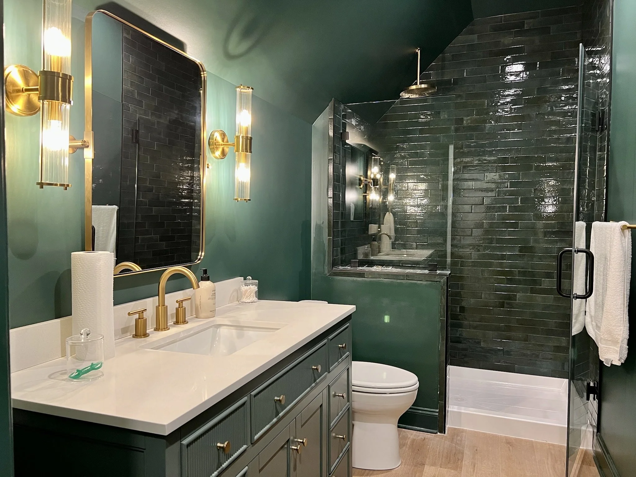 Modern bathroom featuring green walls, a white vanity with gold fixtures, a large mirror flanked by gold lights, a black-tiled shower with glass door, towel rack, bathtub, and wooden floor.