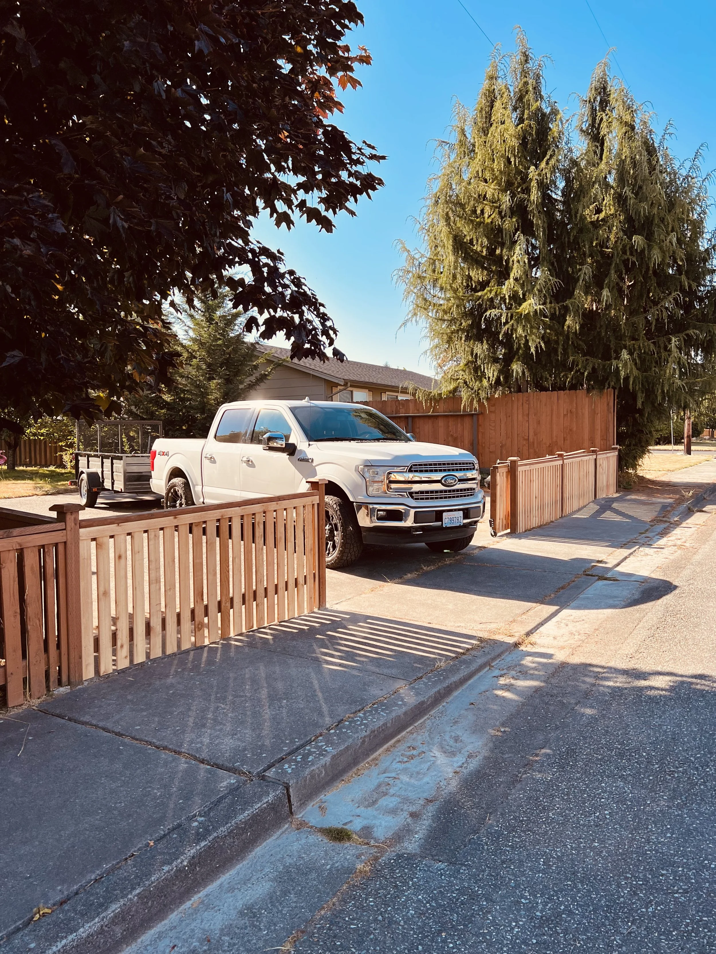 A white pickup truck parked in a residential driveway surrounded by a wooden fence and tall trees, with a clear blue sky overhead.